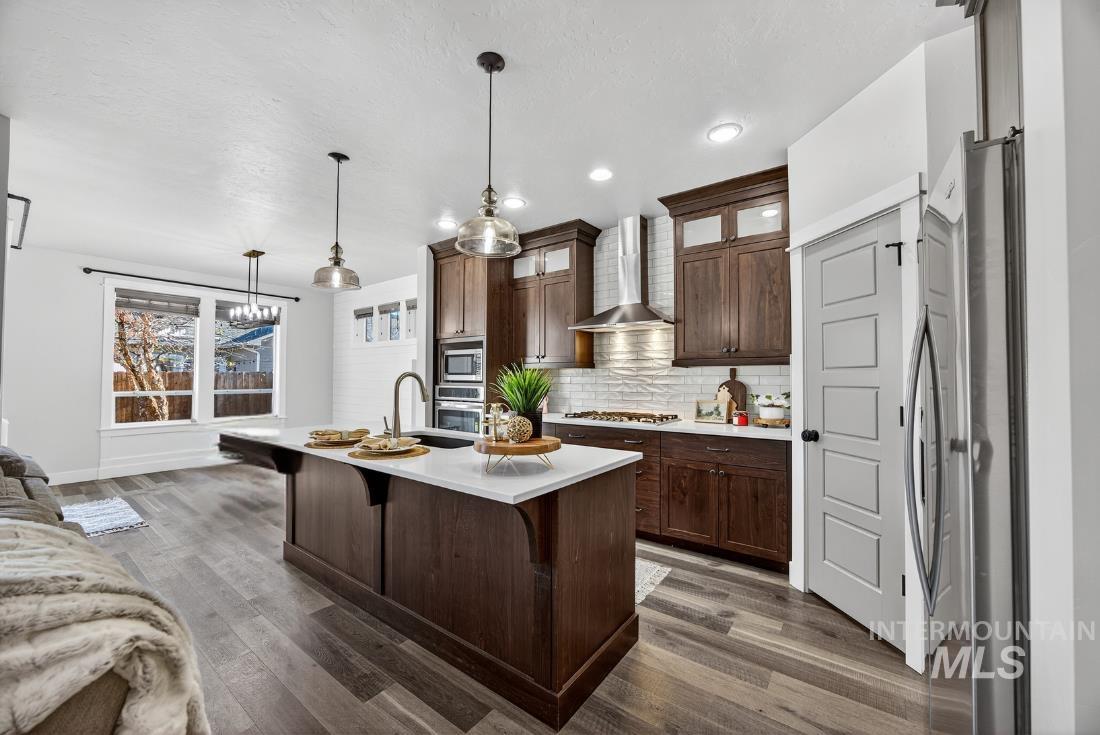 Kitchen with decorative light fixtures, dark brown cabinetry, tasteful backsplash, wall chimney exhaust hood, and an island with sink