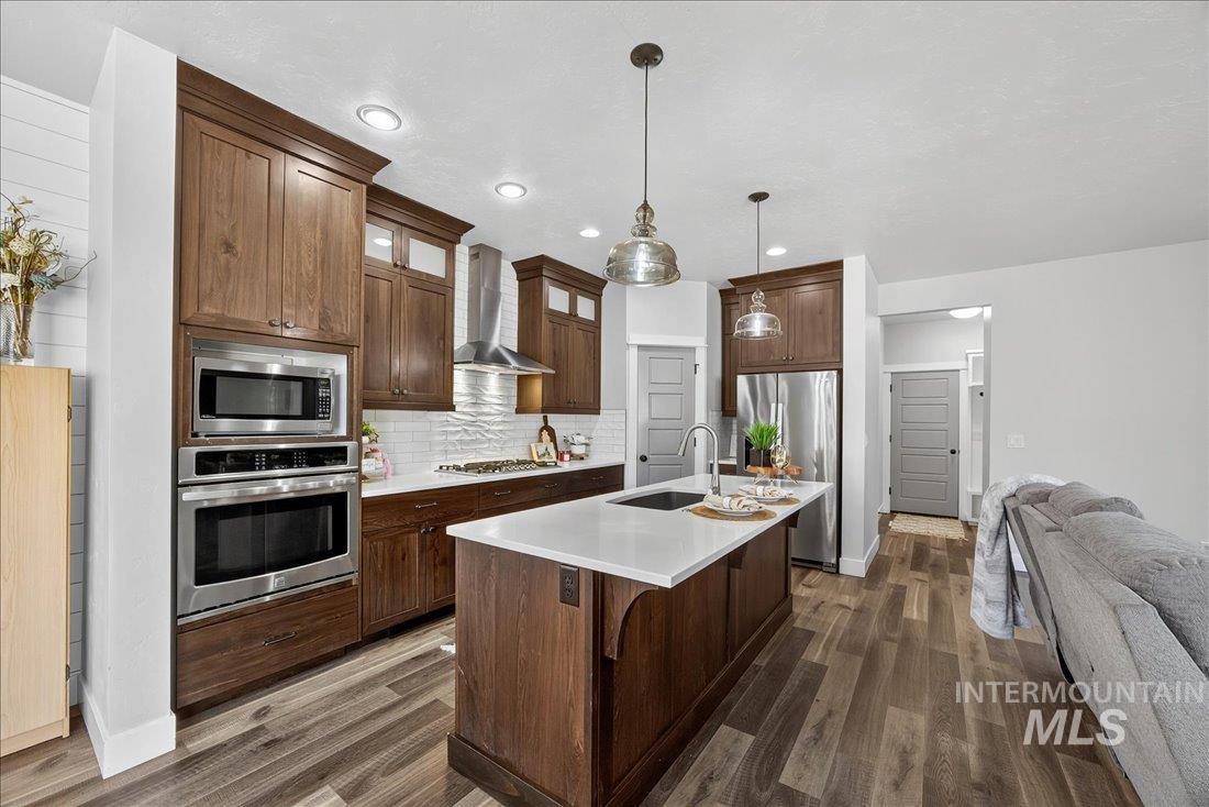 Kitchen with appliances with stainless steel finishes, a kitchen island with sink, hanging light fixtures, wall chimney exhaust hood, and dark wood-type flooring