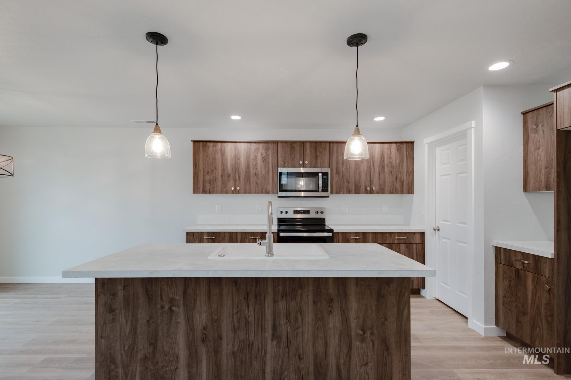 Kitchen with hanging light fixtures, an island with sink, stainless steel appliances, brown cabinets, and light wood-style flooring