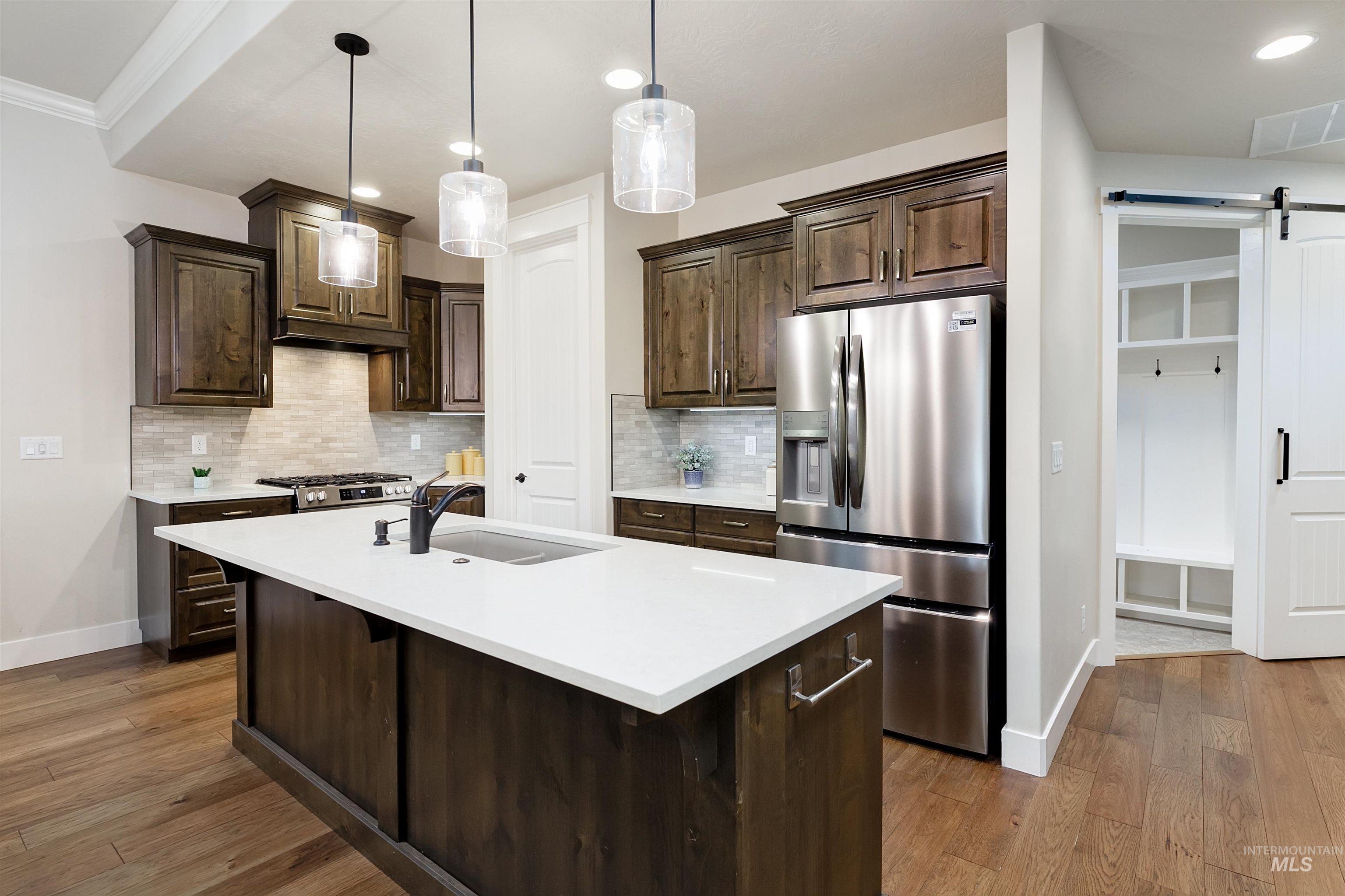 Kitchen featuring a barn door, appliances with stainless steel finishes, dark brown cabinets, decorative backsplash, and a kitchen island with sink