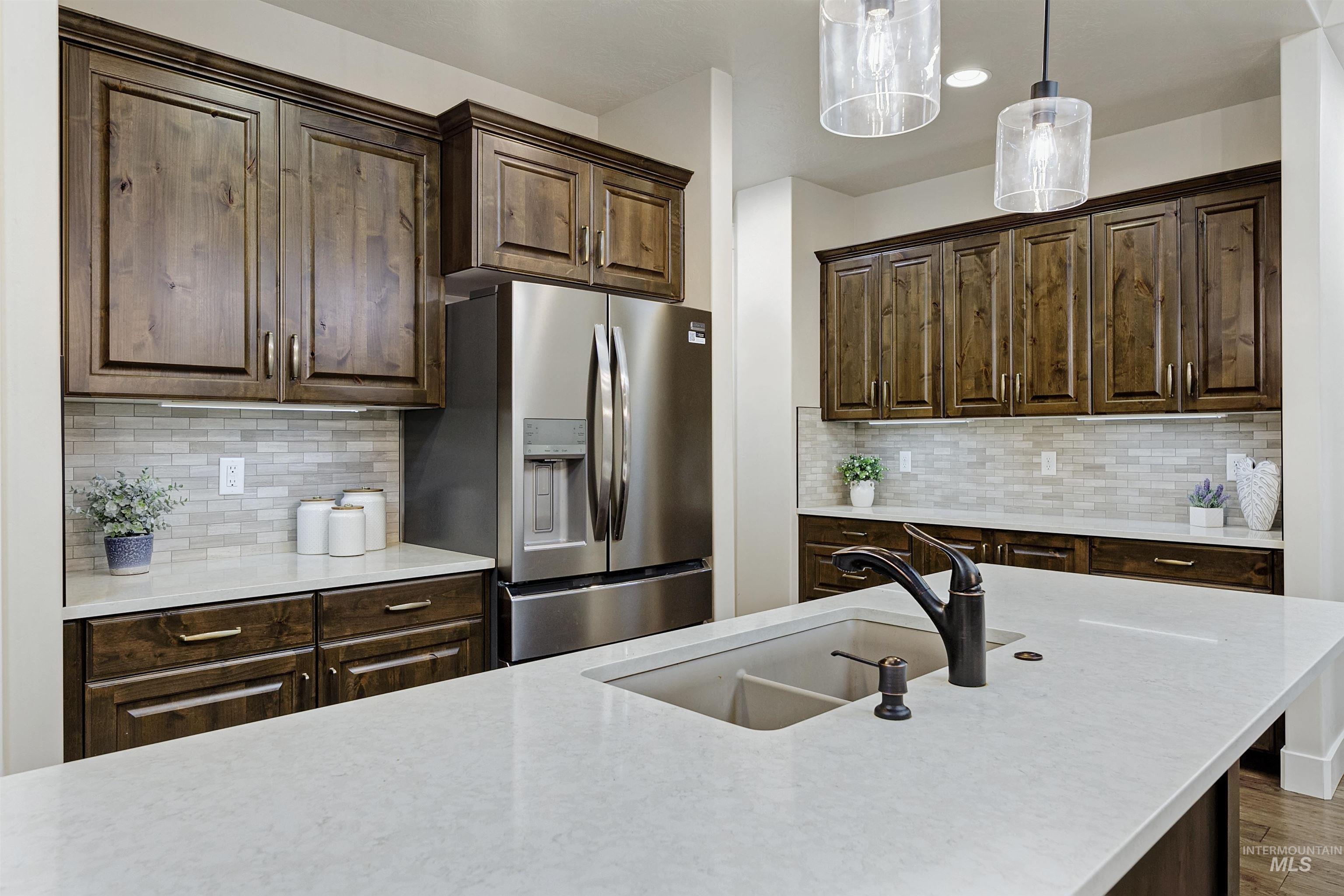 Kitchen with dark brown cabinets, stainless steel fridge, hanging light fixtures, light stone counters, and decorative backsplash