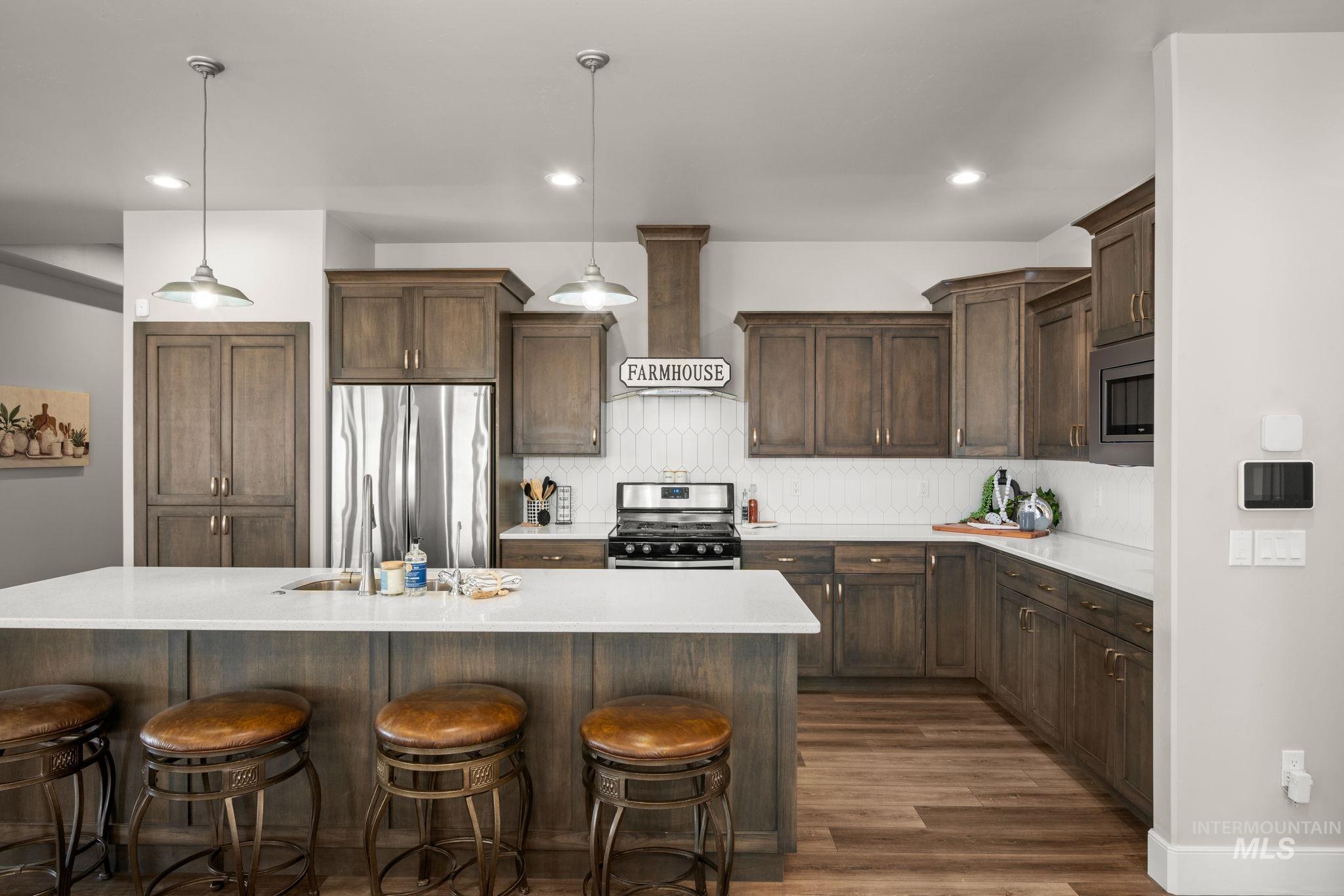 Kitchen featuring decorative light fixtures, appliances with stainless steel finishes, wall chimney exhaust hood, an island with sink, and backsplash