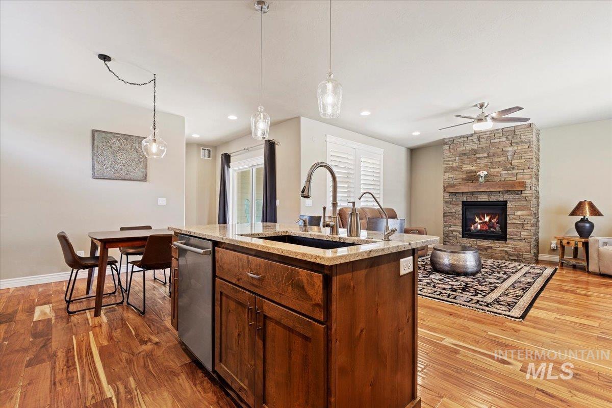 Kitchen featuring a ceiling fan, wood finished floors, stainless steel dishwasher, a center island with sink, and light stone counters