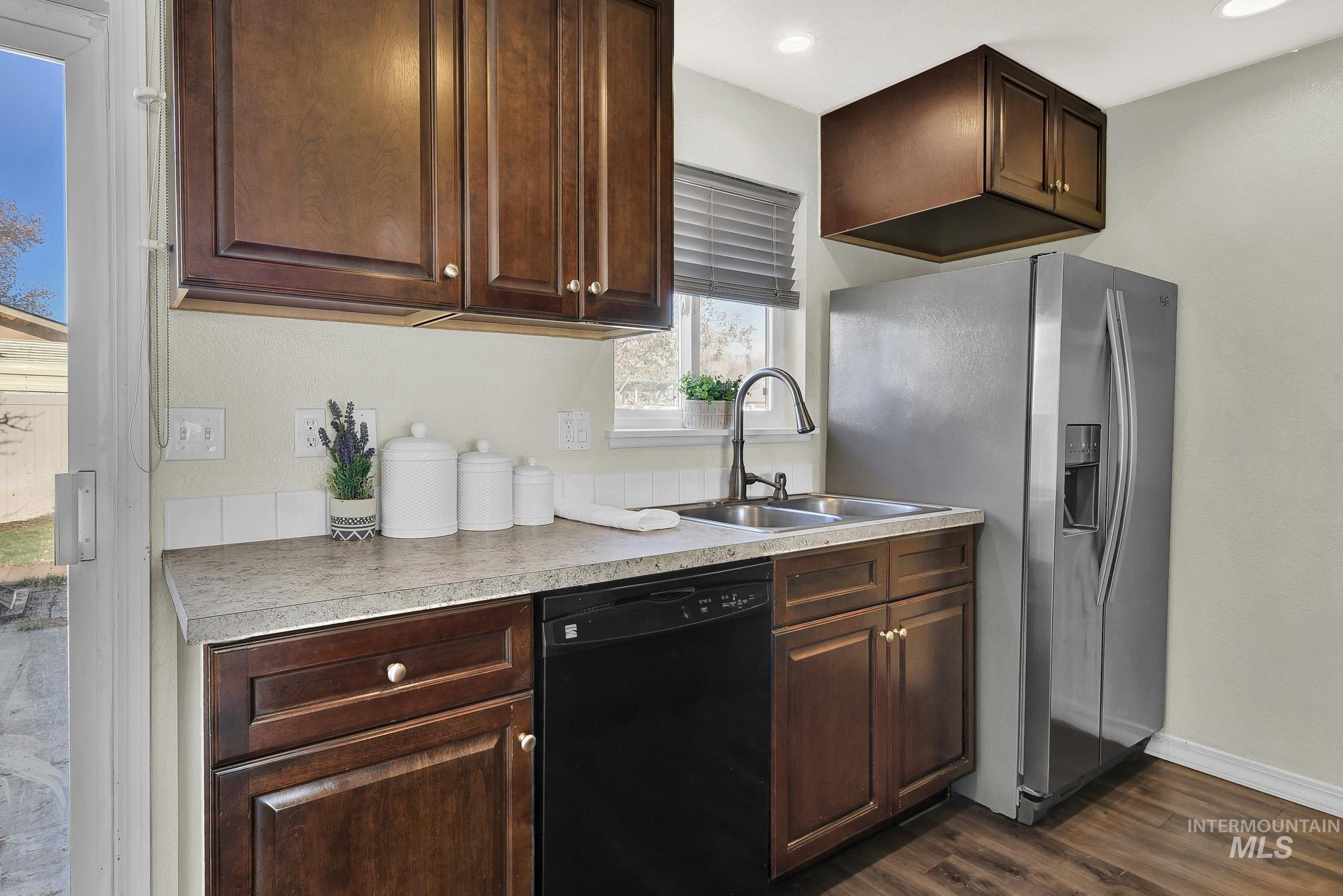 Kitchen with black dishwasher, rich brown cabinets, stainless steel refrigerator with ice dispenser, laminate wood-style flooring, and neutral granite style countertops