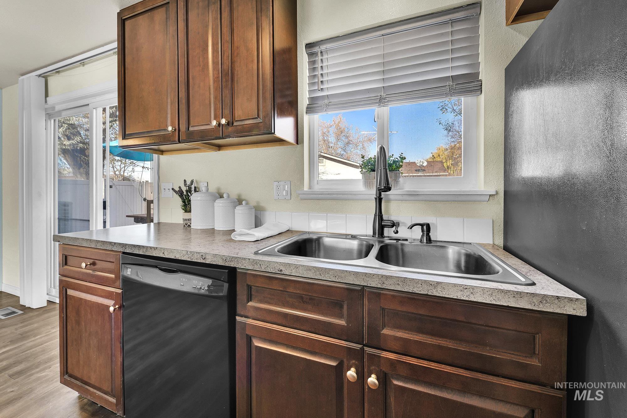 Kitchen with black dishwasher, light granite look countertops, stainless steel sink with window above, laminate wood-style flooring, and plenty of natural light