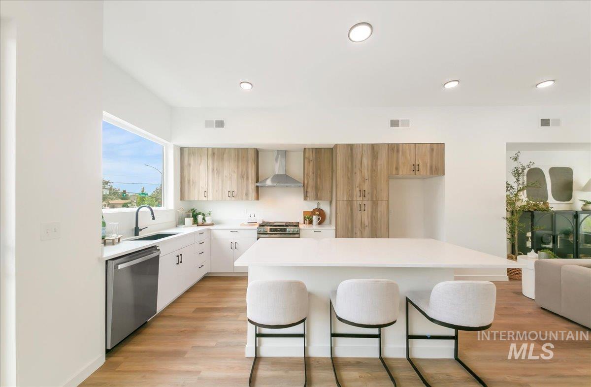 Kitchen featuring a kitchen breakfast bar, light countertops, light wood-style flooring, stainless steel appliances, and wall chimney range hood