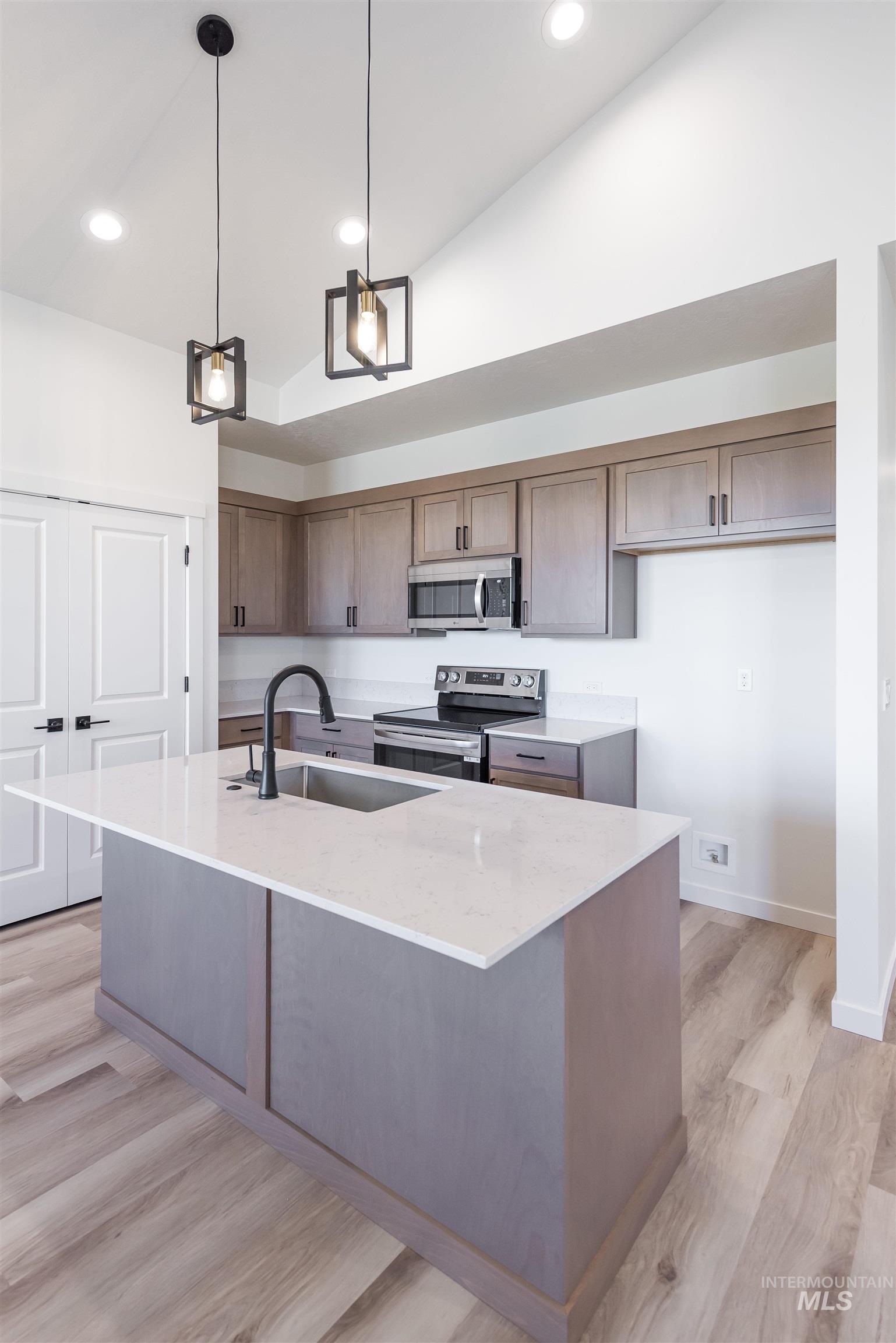 Kitchen with light stone counters, decorative light fixtures, a kitchen island with sink, appliances with stainless steel finishes, and light wood finished floors