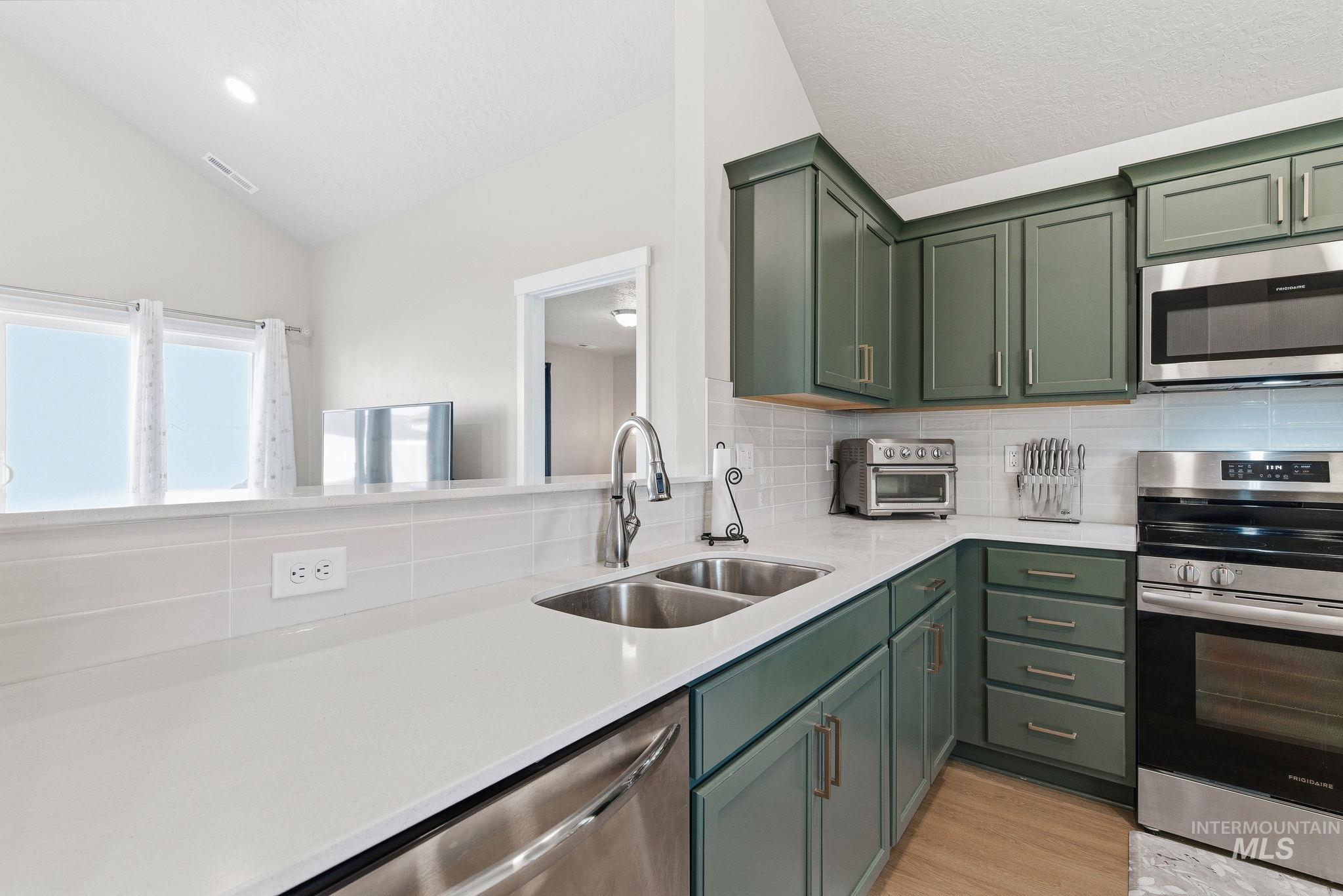 Kitchen featuring green cabinetry, appliances with stainless steel finishes, lofted ceiling, light stone countertops, and tasteful backsplash