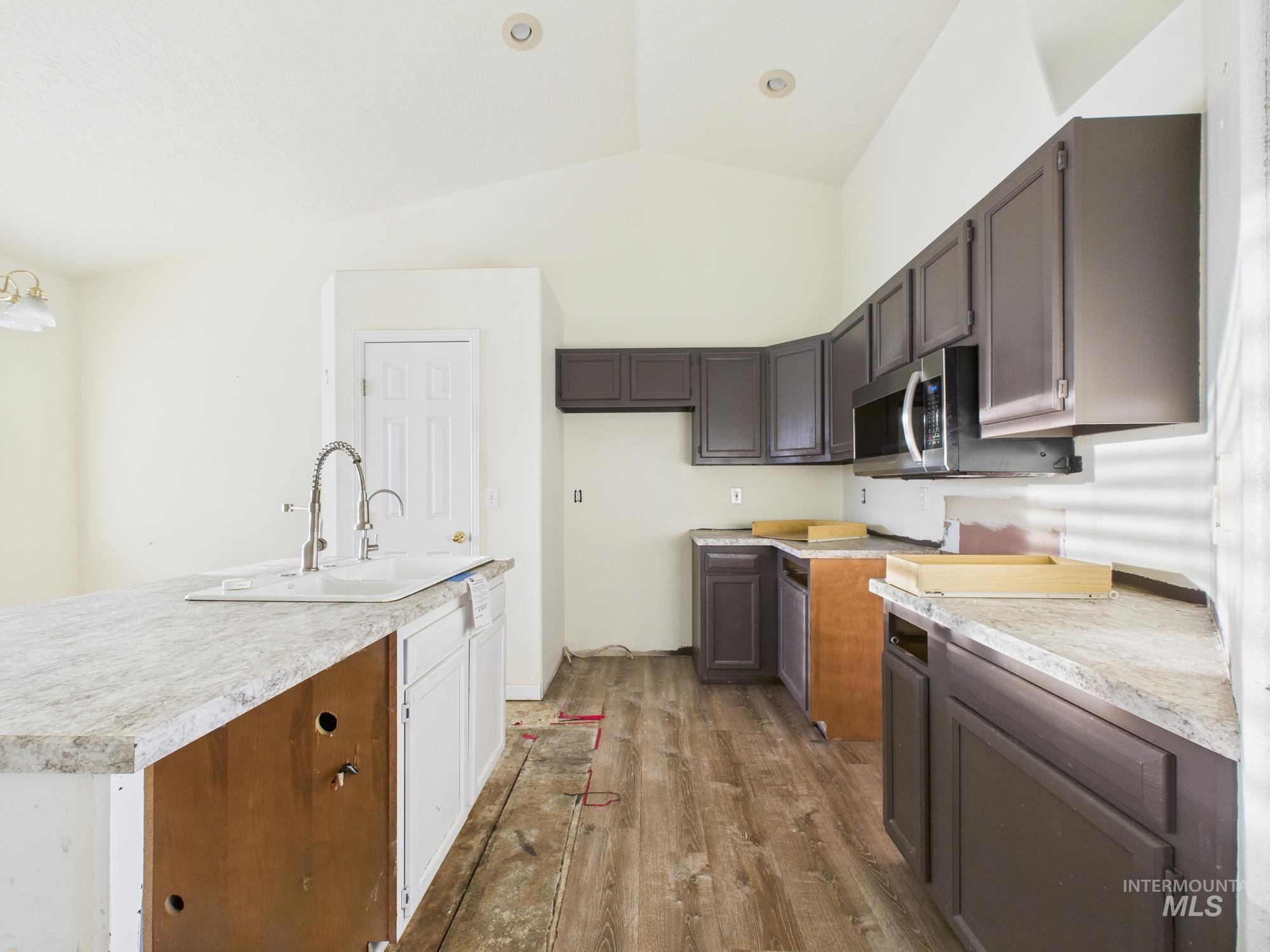 Kitchen featuring light countertops, wood finished floors, stainless steel microwave, vaulted ceiling, and a kitchen island with sink