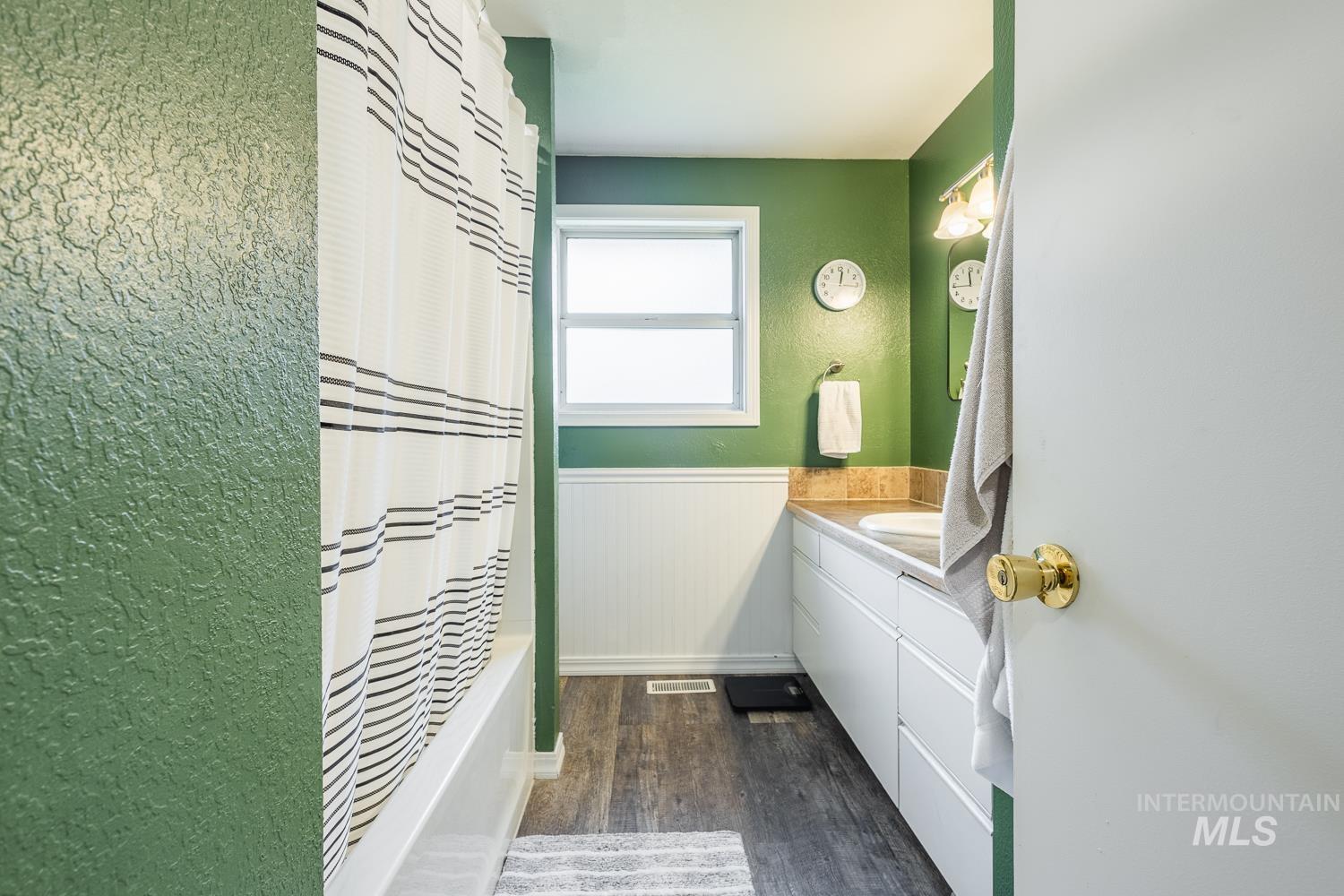 Bathroom featuring vanity, shower / bathtub combination with curtain, a textured wall, dark wood-type flooring, and a wainscoted wall