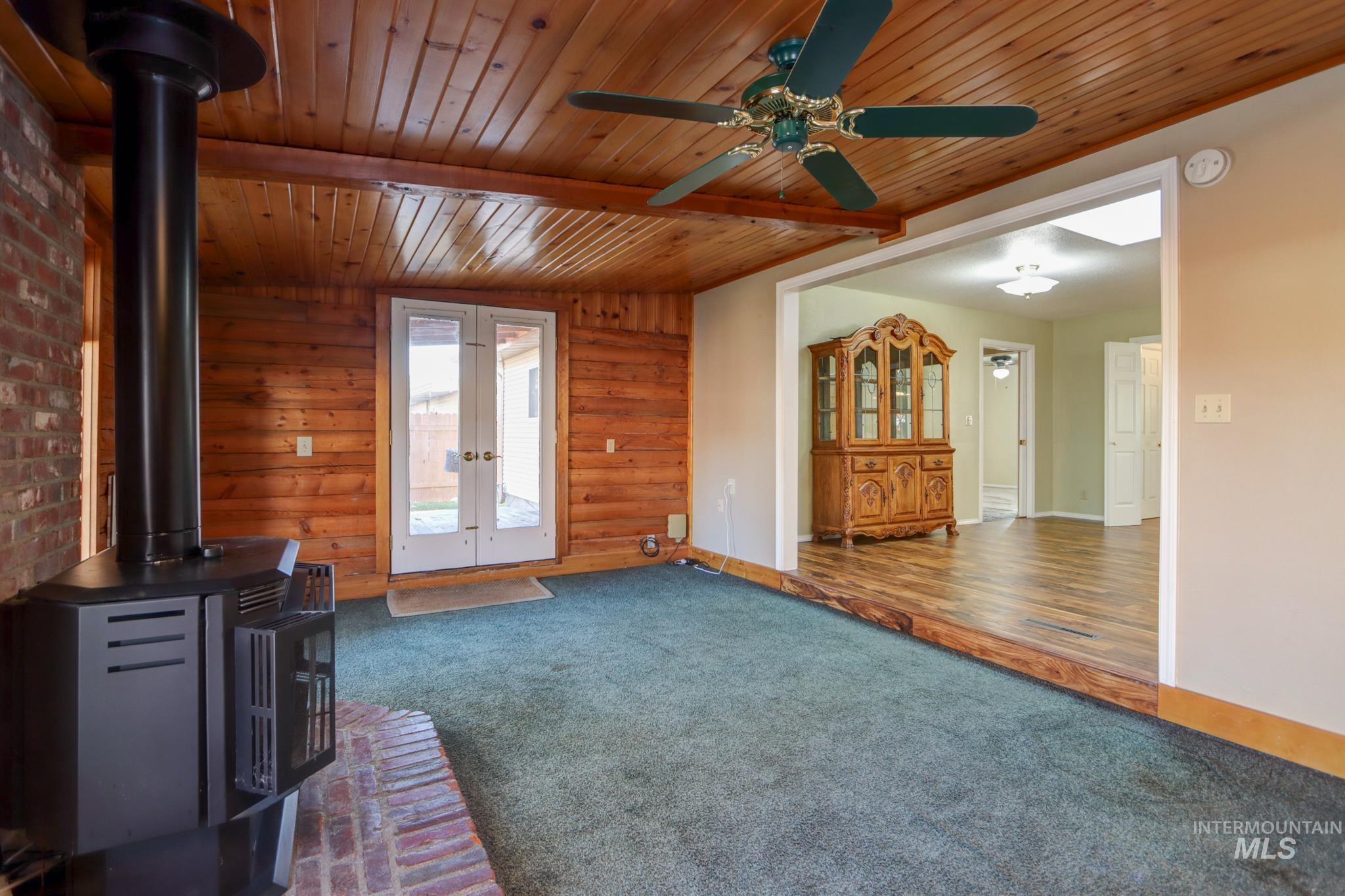 Unfurnished living room featuring a wood stove, carpet, wood walls, a wooden ceiling with exposed beams, and french doors