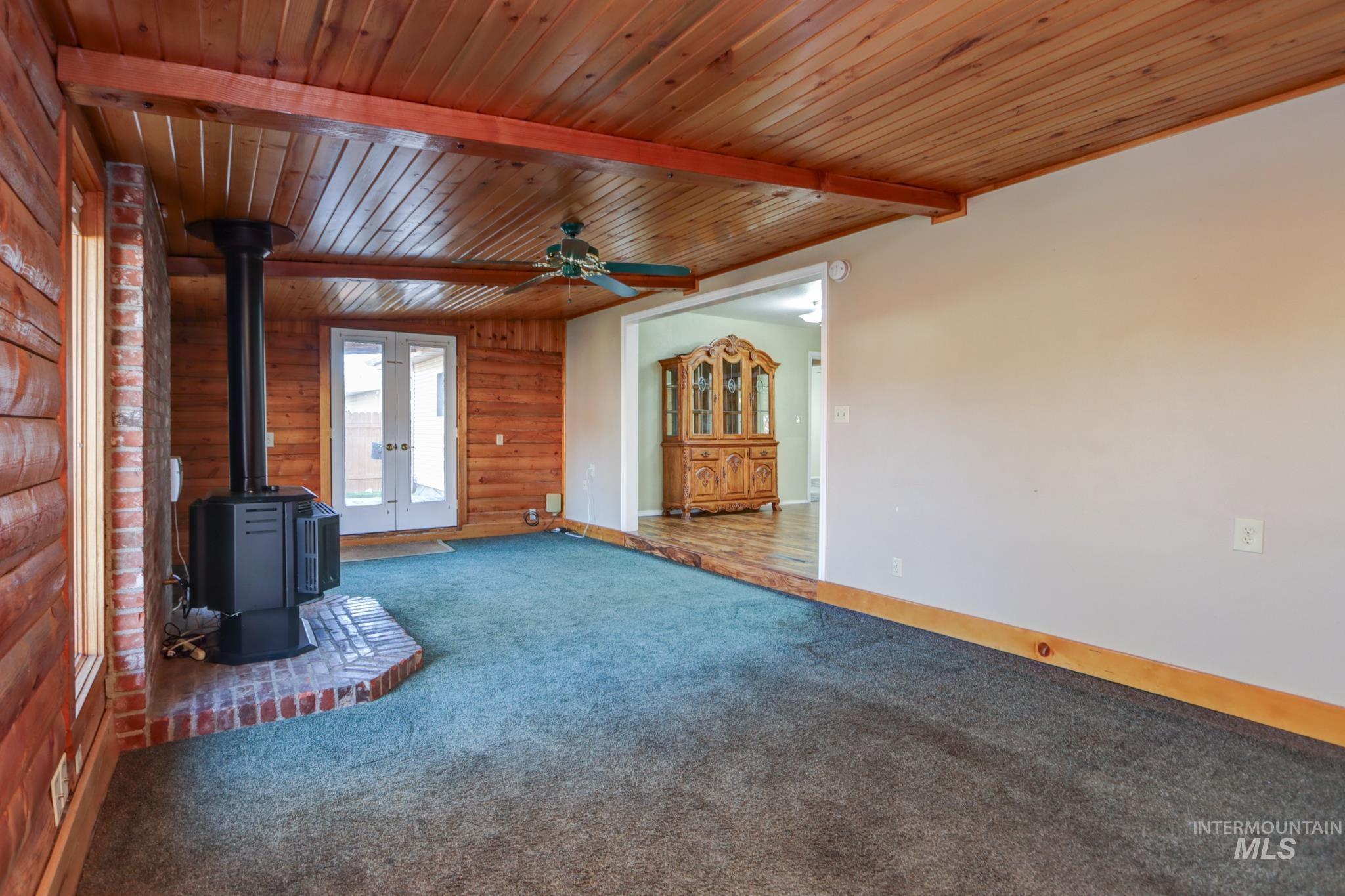 Unfurnished living room with a wood stove, wooden walls, carpet floors, a wooden ceiling with exposed beams, and ceiling fan