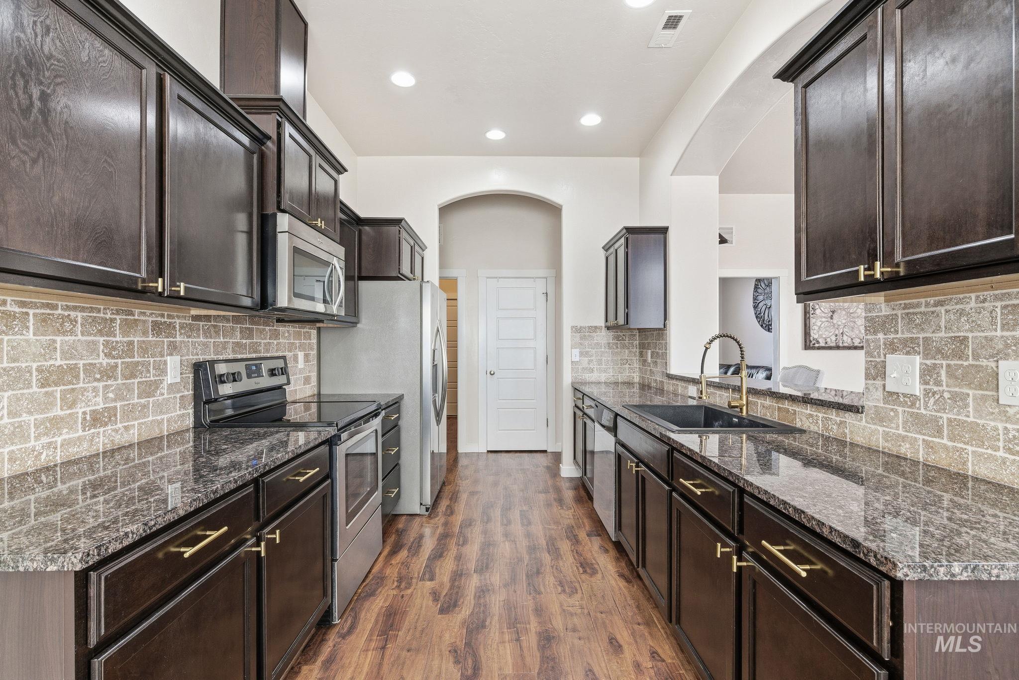 Kitchen featuring stainless steel appliances, dark stone countertops, dark wood-type flooring, dark brown cabinets, and tasteful backsplash