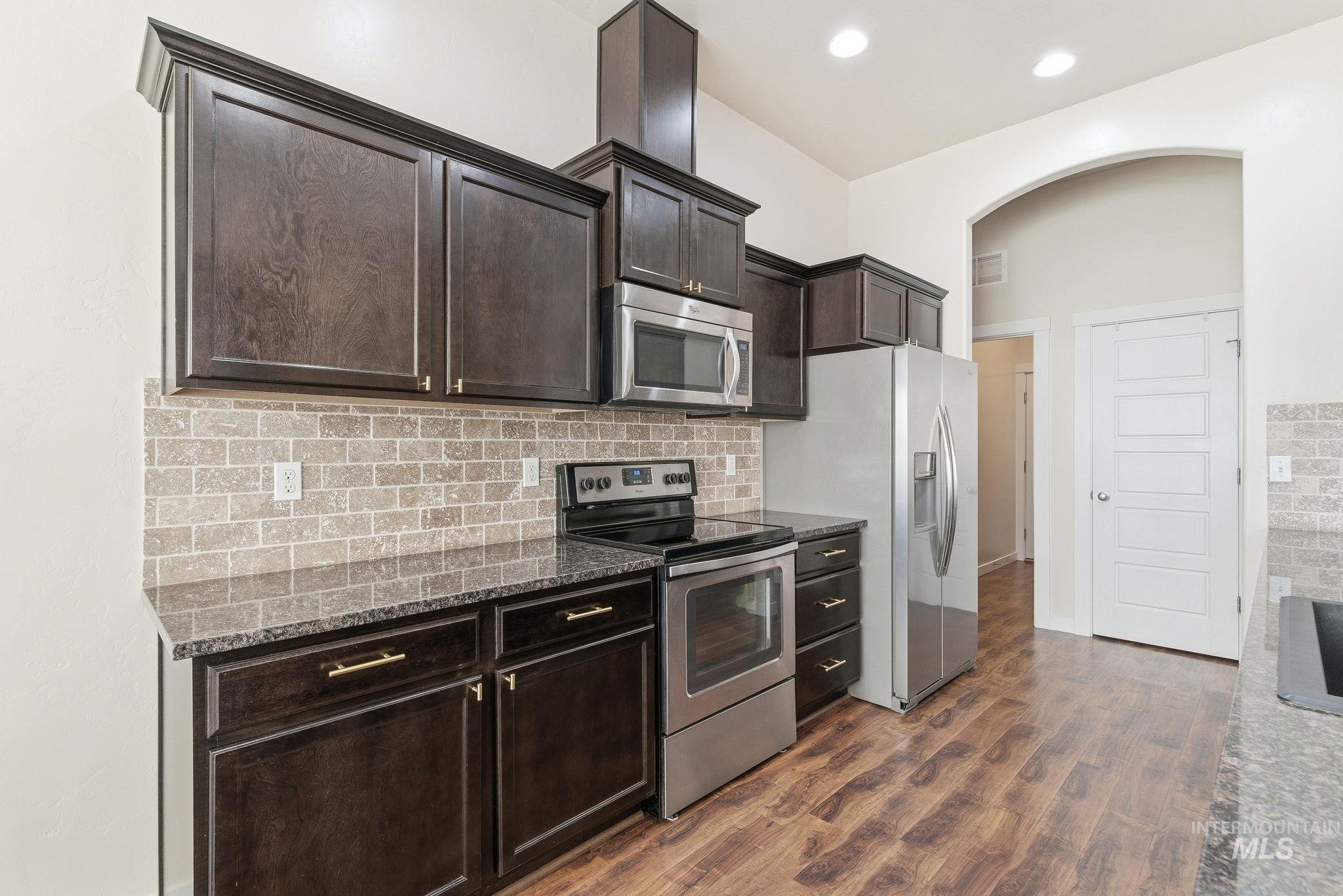 Kitchen with stainless steel appliances, dark stone counters, dark brown cabinets, tasteful backsplash, and dark wood-style flooring