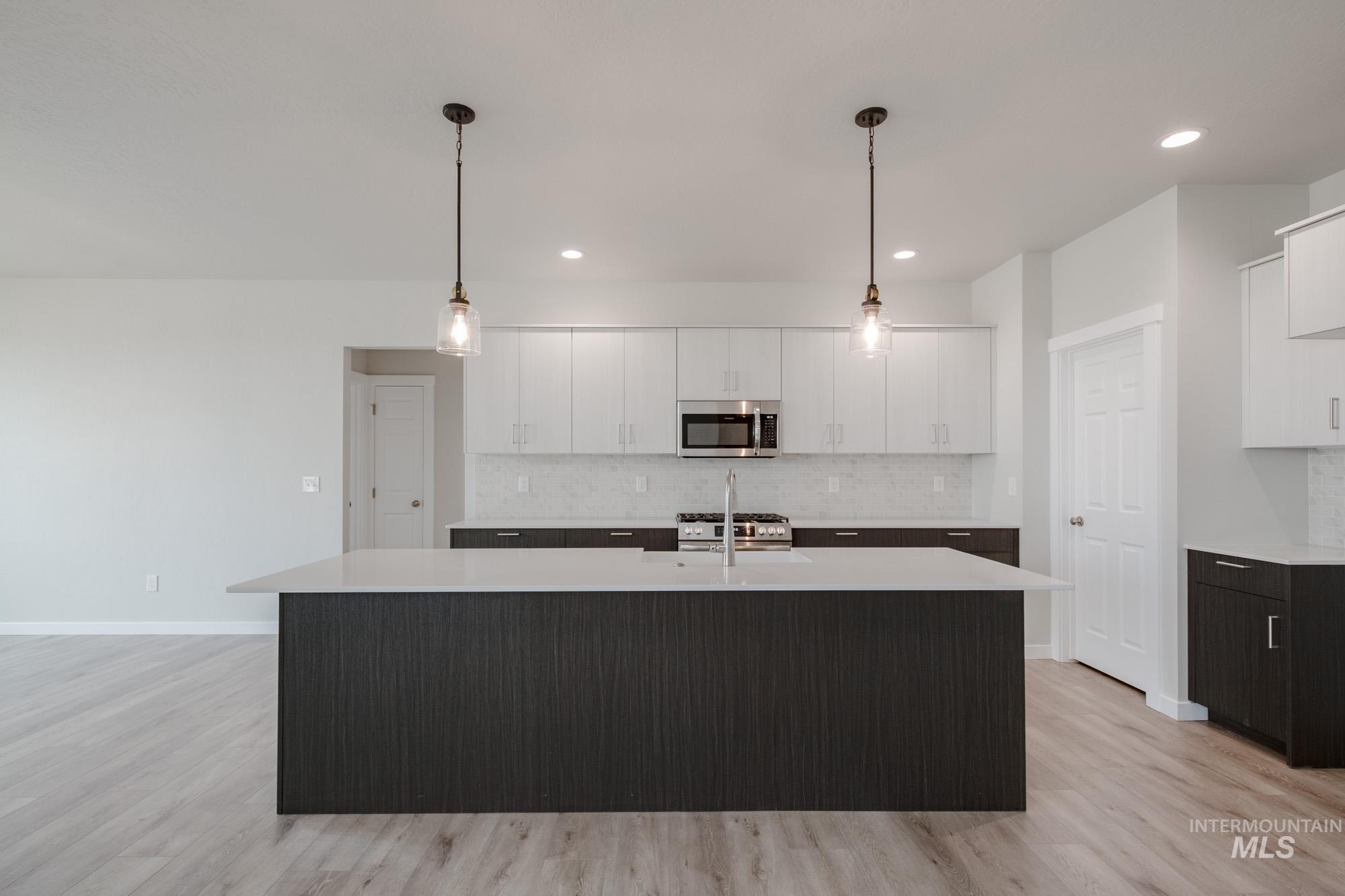 Kitchen featuring white cabinetry, a center island with sink, decorative light fixtures, stainless steel microwave, and backsplash