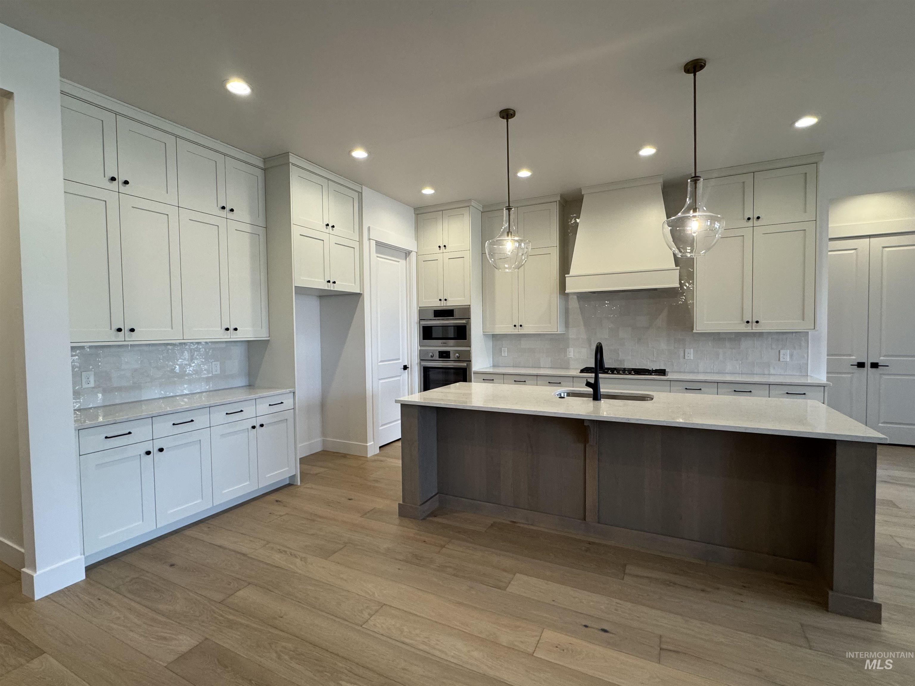 Kitchen with decorative light fixtures, light stone counters, white cabinetry, custom range hood, and a center island with sink