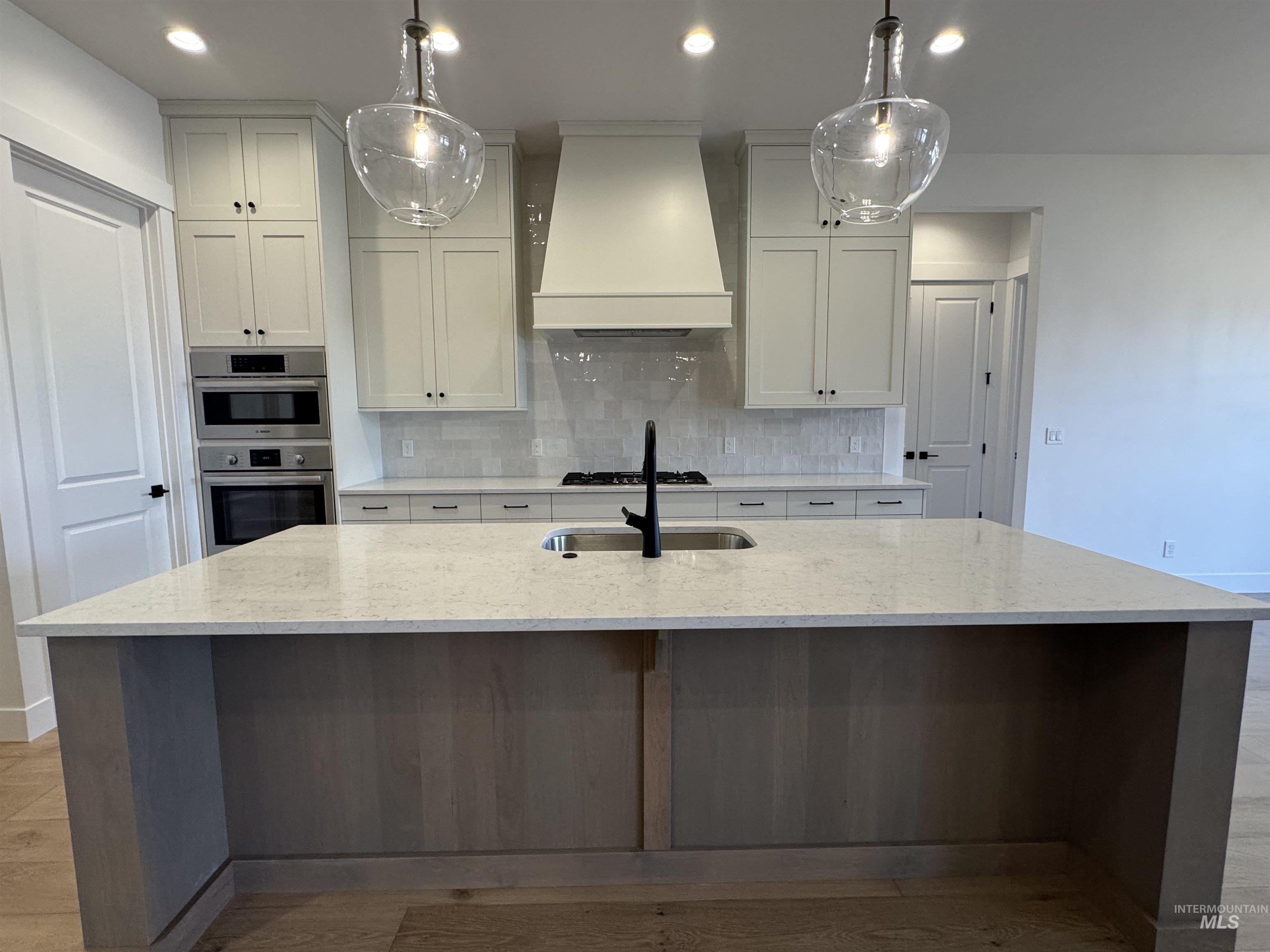 Kitchen featuring custom range hood, tasteful backsplash, light stone counters, decorative light fixtures, and a center island with sink