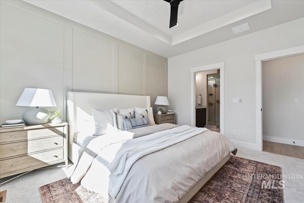 Carpeted bedroom featuring a decorative wall, a raised ceiling, ensuite bathroom, and ceiling fan