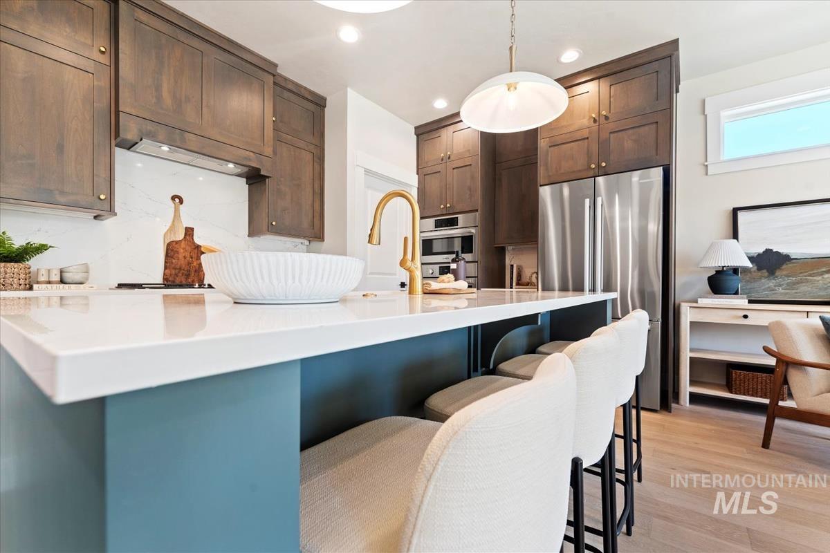 Kitchen featuring stainless steel appliances, pendant lighting, light stone counters, light wood-style floors, and a center island with sink