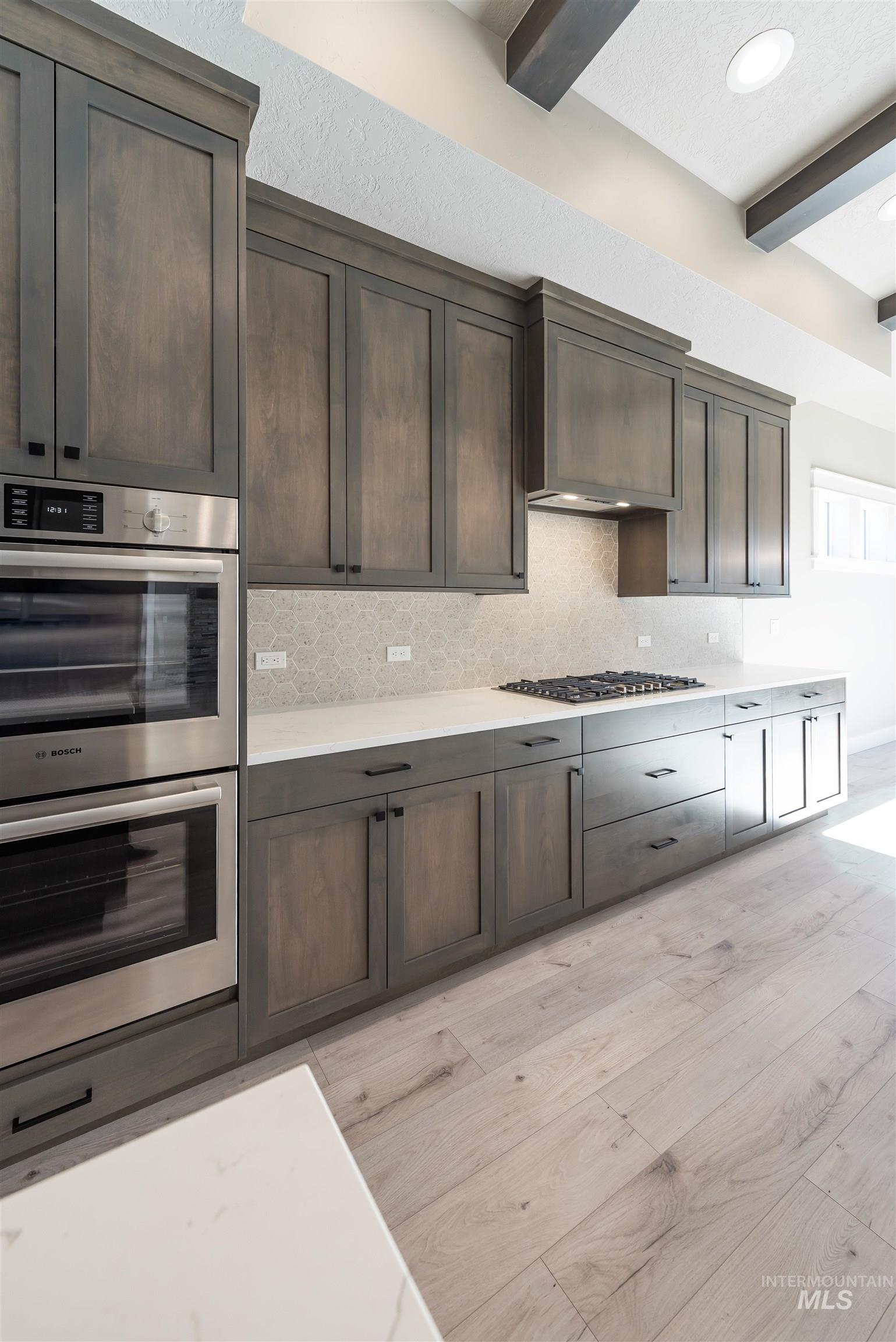 Kitchen featuring stainless steel appliances, beamed ceiling, backsplash, light stone counters, and dark brown cabinetry