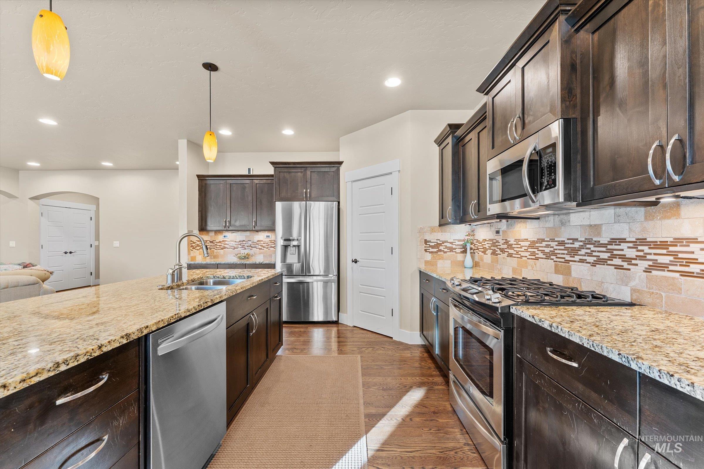 Kitchen with dark brown cabinets, stainless steel appliances, arched walkways, light stone countertops, and hanging light fixtures