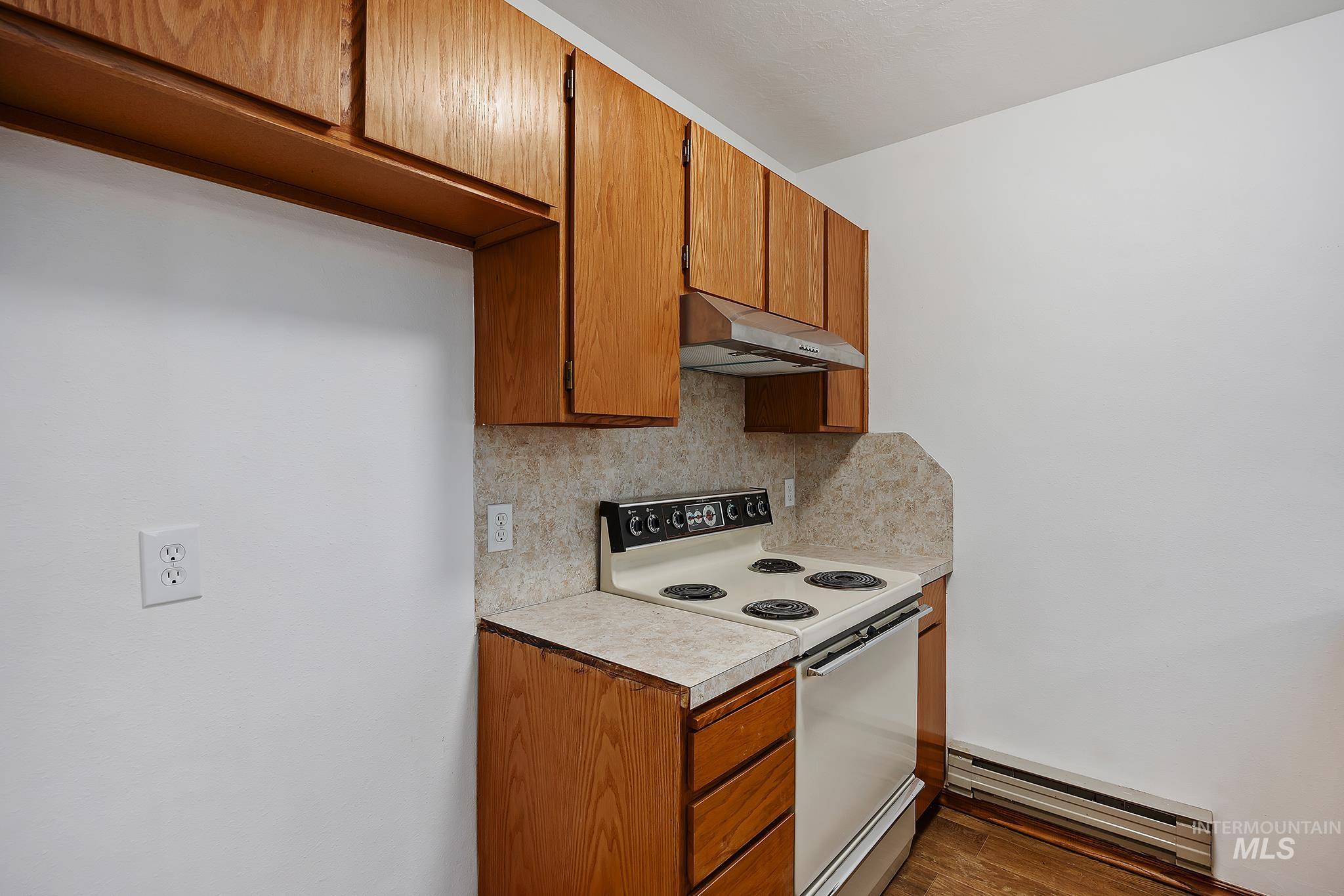 Kitchen featuring electric stove, brown cabinets, baseboard heating, light countertops, and under cabinet range hood