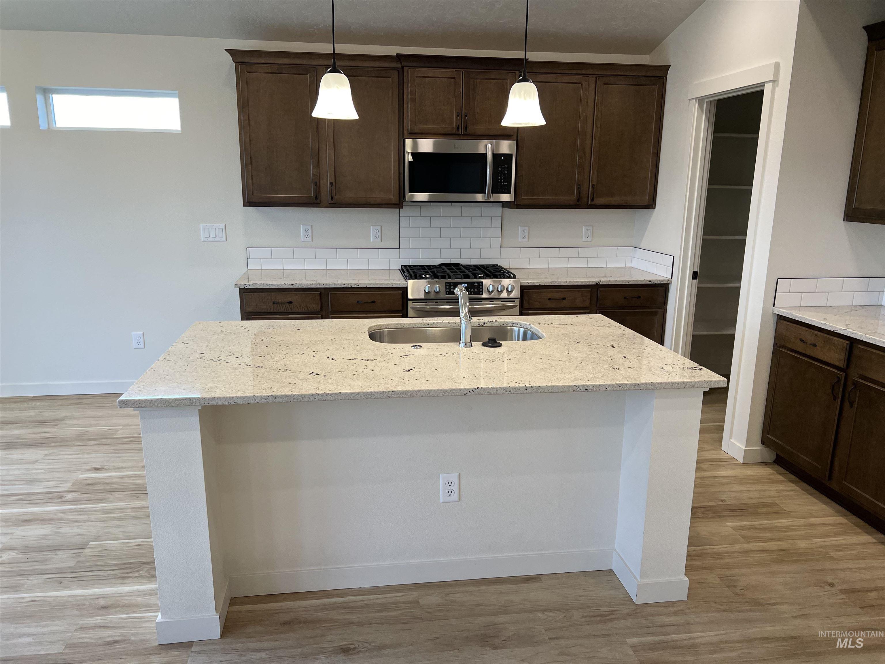 Kitchen featuring an island with sink, decorative light fixtures, stainless steel appliances, dark brown cabinets, and light stone countertops