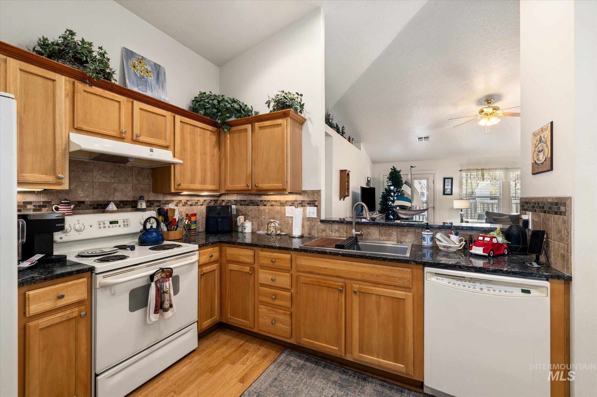 Kitchen with white appliances, lofted ceiling, brown cabinets, under cabinet range hood, and backsplash