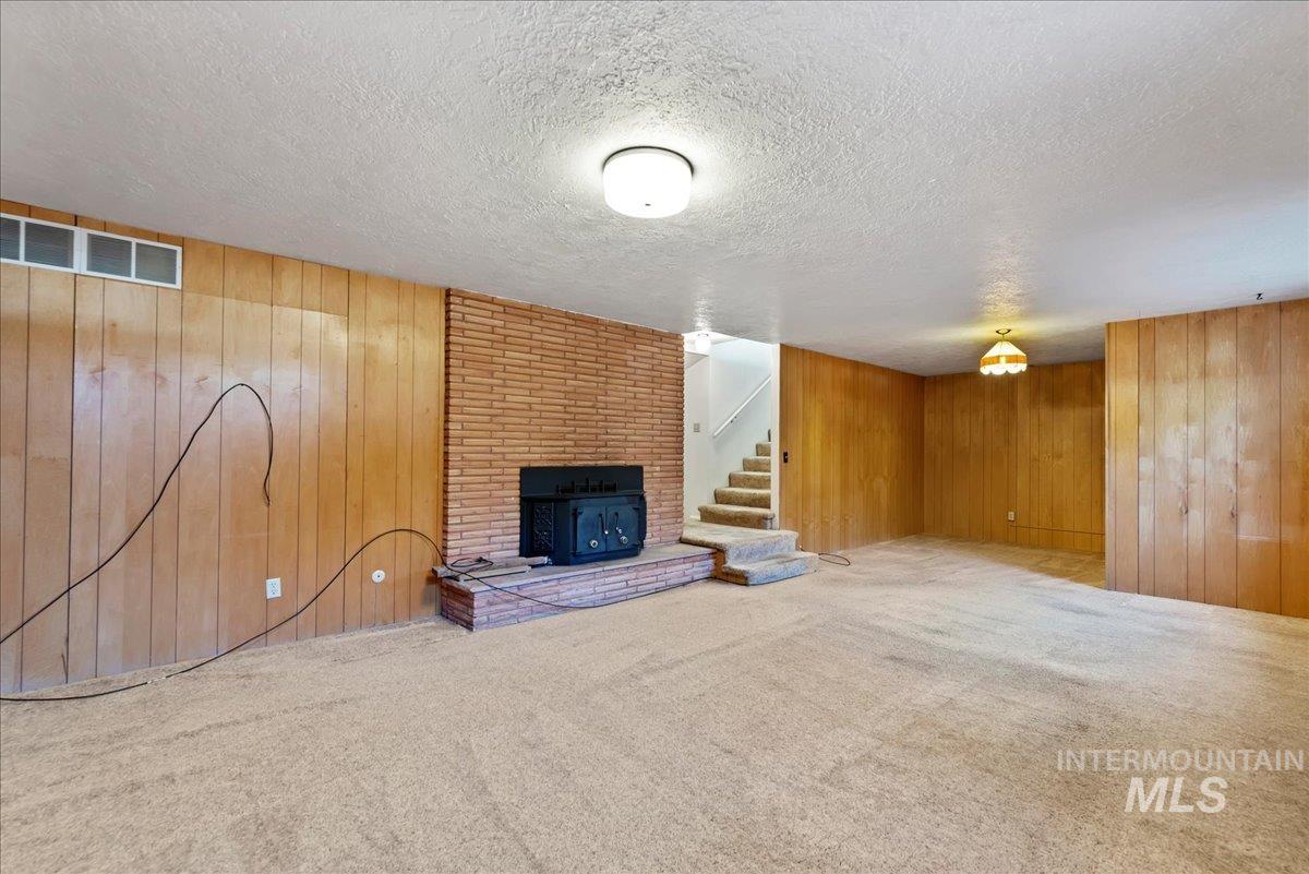 Unfurnished living room with a textured ceiling, stairs, wooden walls, a brick fireplace, and carpet flooring