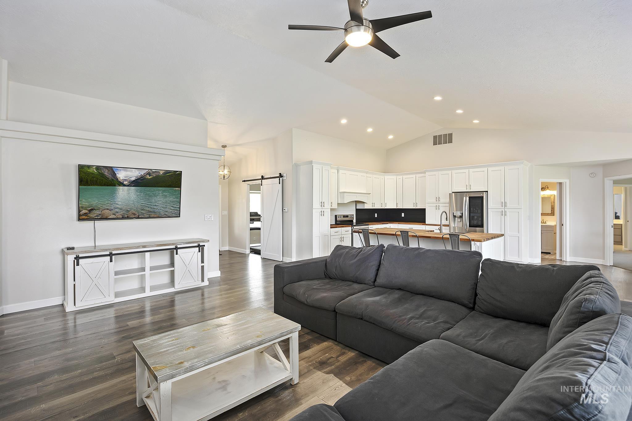Living room with a barn door, vaulted ceiling, dark wood-style flooring, and ceiling fan
