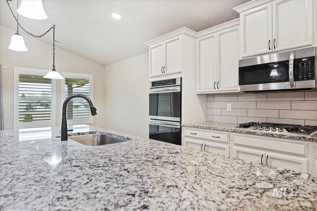 Kitchen with backsplash, light stone counters, stainless steel appliances, pendant lighting, and white cabinetry