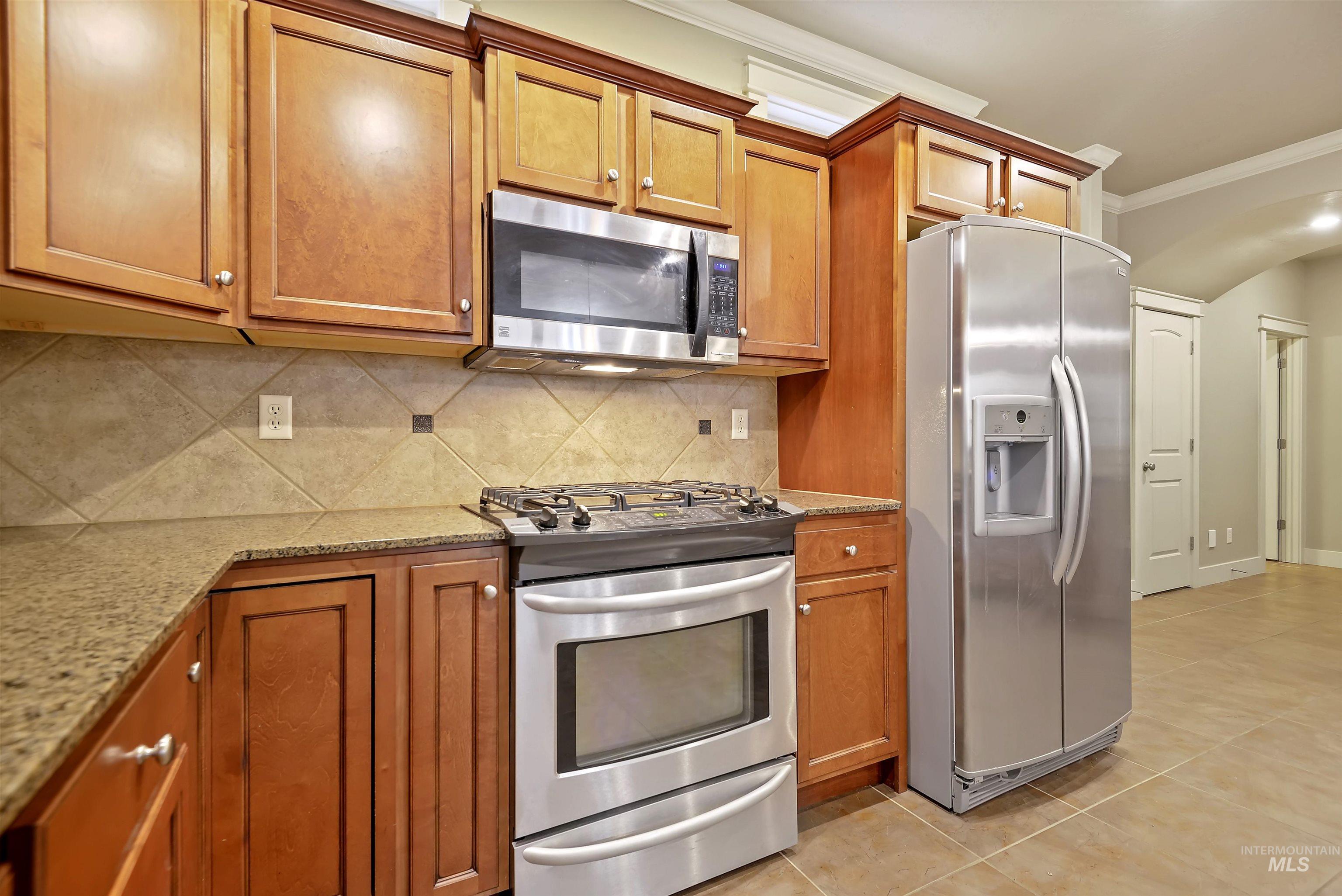 Kitchen featuring stainless steel appliances, brown cabinets, arched walkways, light stone counters, and crown molding