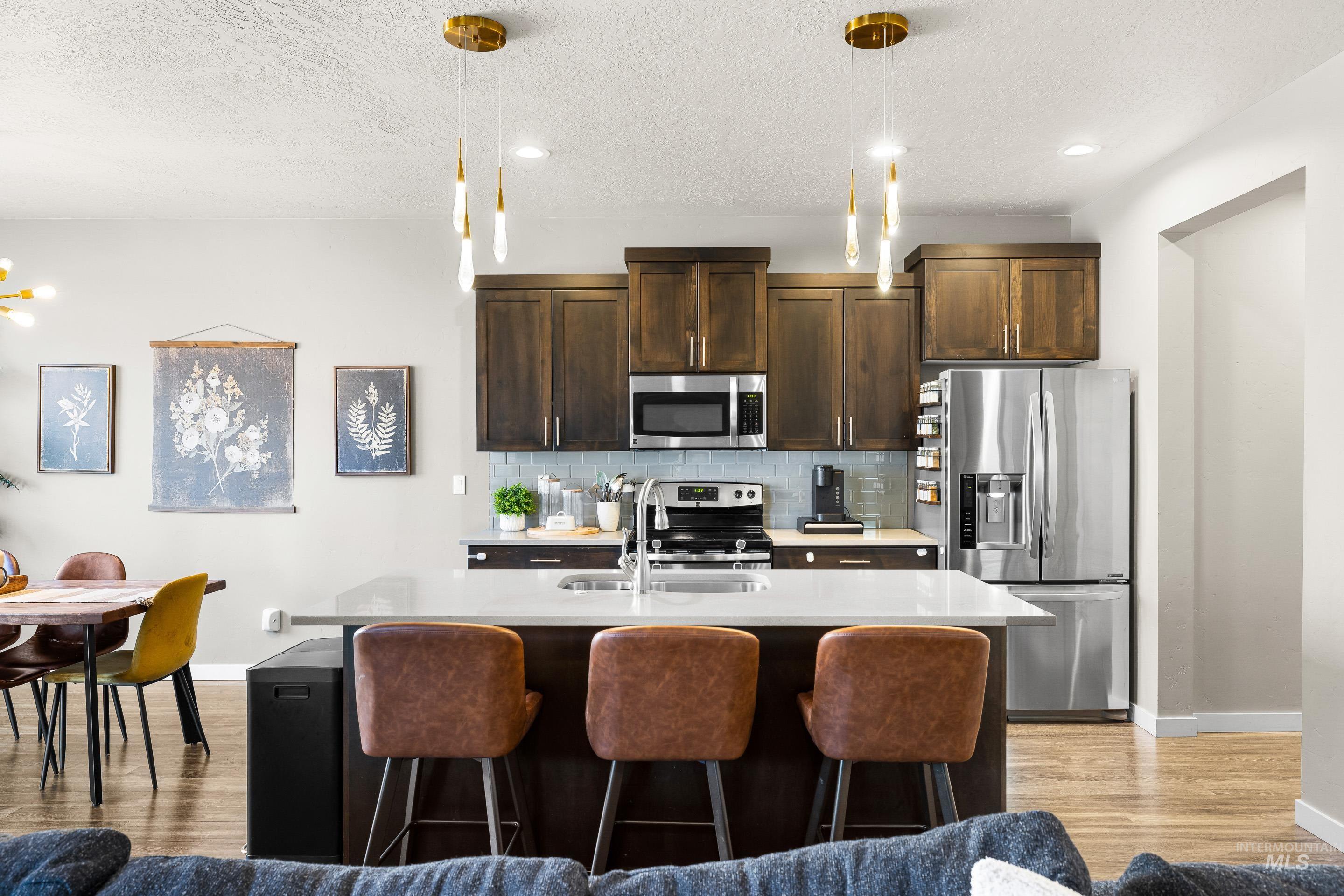 Kitchen featuring appliances with stainless steel finishes, dark brown cabinets, decorative light fixtures, a kitchen island with sink, and tasteful backsplash