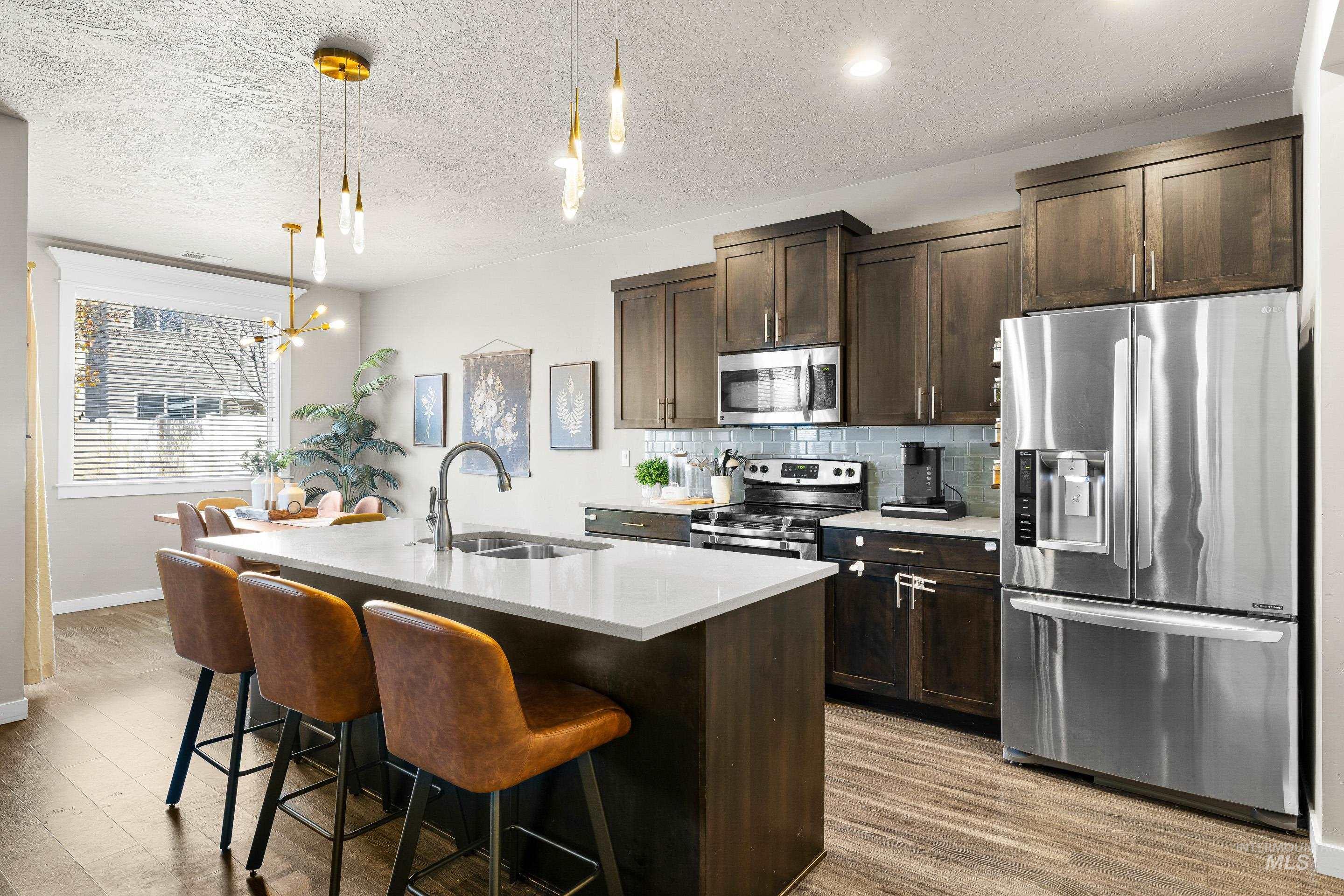 Kitchen featuring stainless steel appliances, dark brown cabinets, pendant lighting, a textured ceiling, and a kitchen island with sink