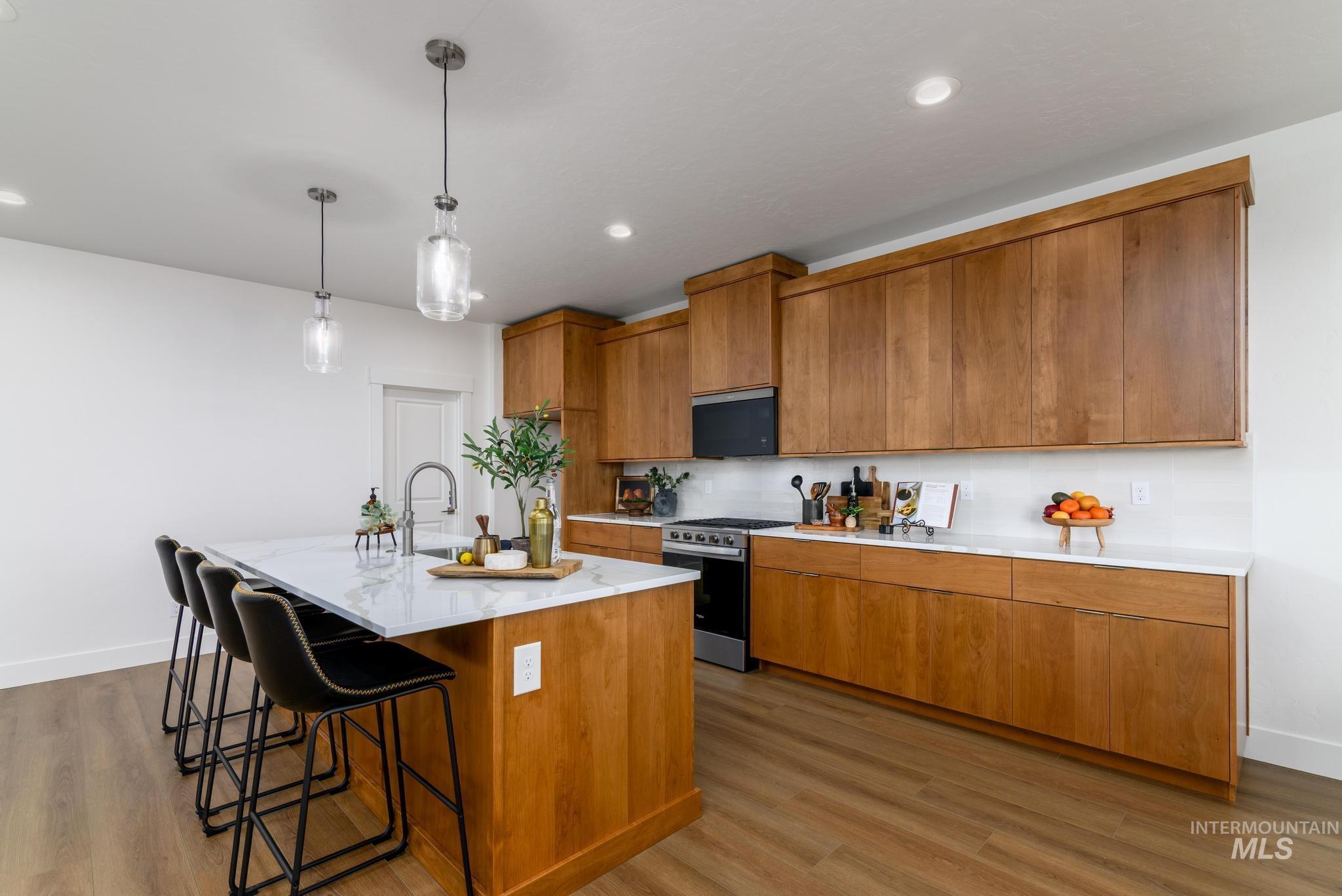 Kitchen with brown cabinets, a center island with sink, a kitchen breakfast bar, stainless steel gas stove, and decorative light fixtures