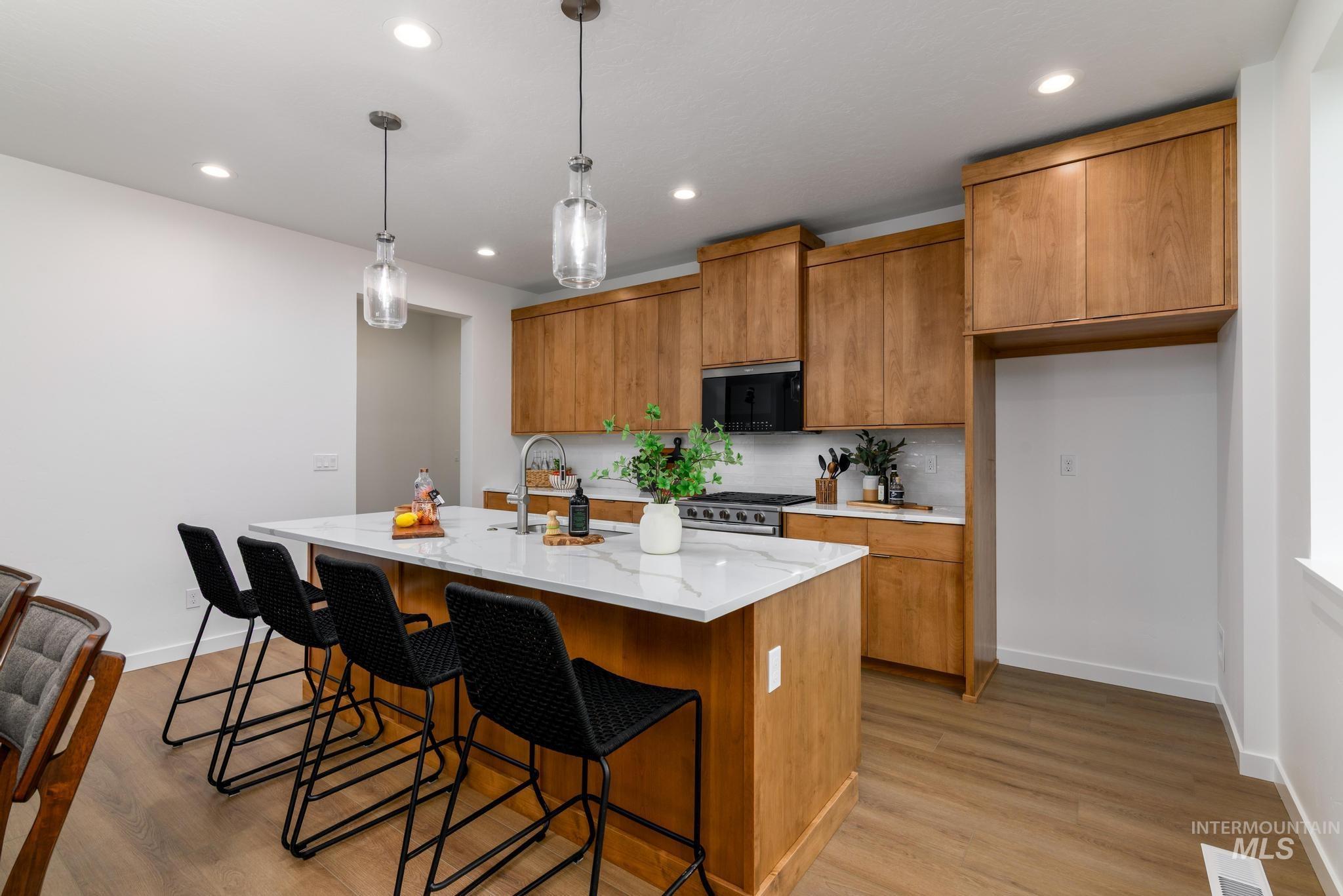Kitchen with brown cabinets, an island with sink, light wood-style flooring, hanging light fixtures, and light stone countertops