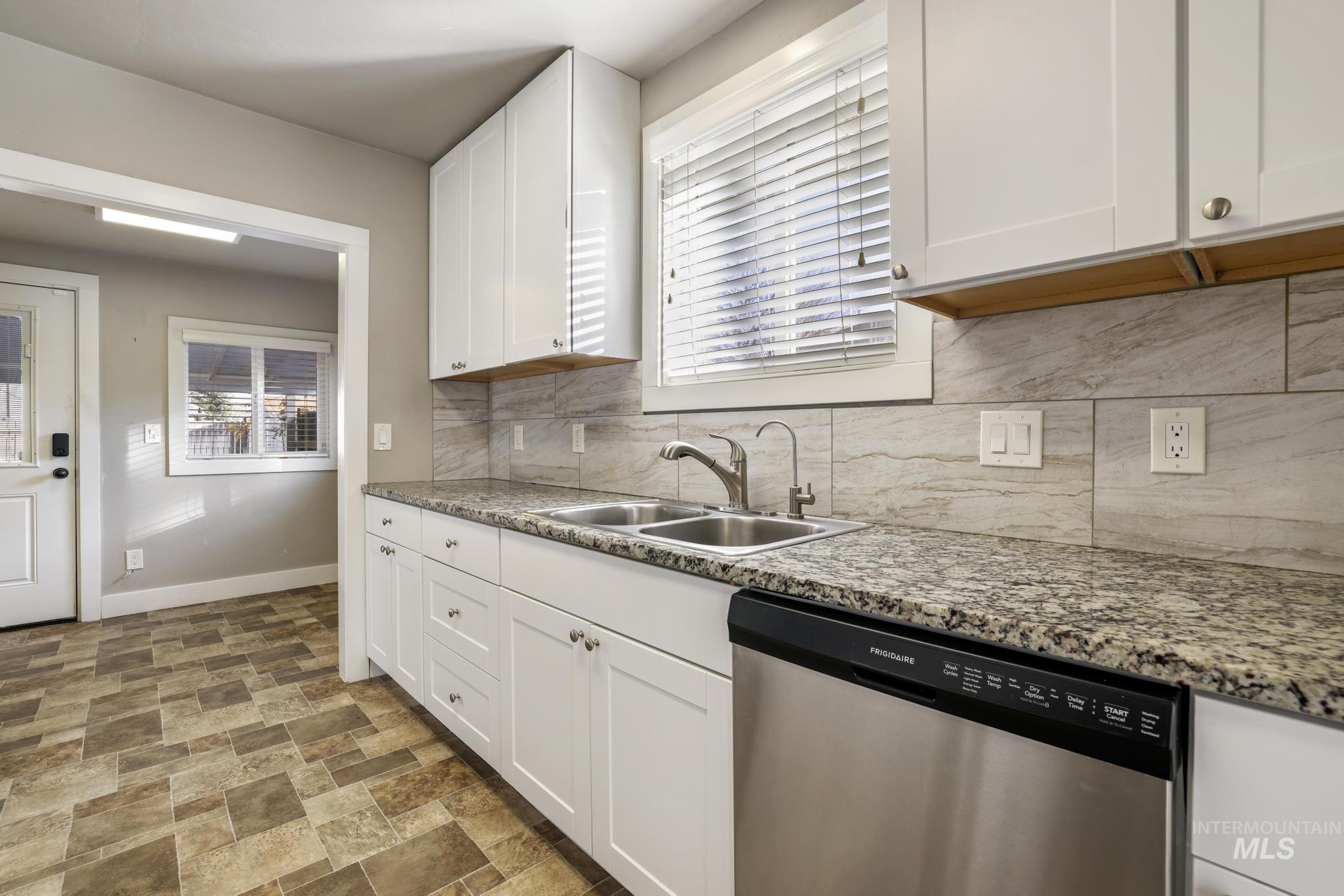 Kitchen with stainless steel dishwasher, tasteful backsplash, white cabinets, stone finish flooring, and light stone counters