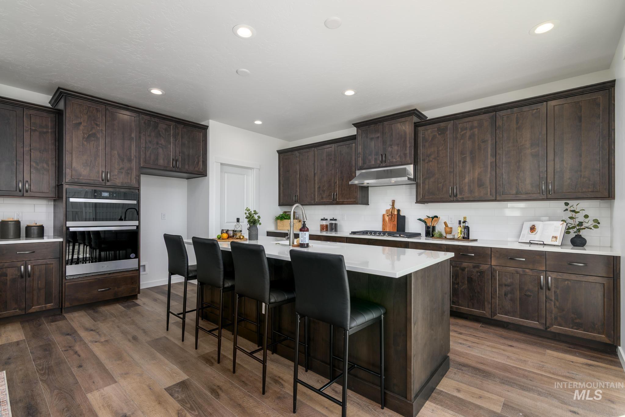 Kitchen with tasteful backsplash, dark brown cabinetry, stainless steel double oven, a kitchen island with sink, and a breakfast bar