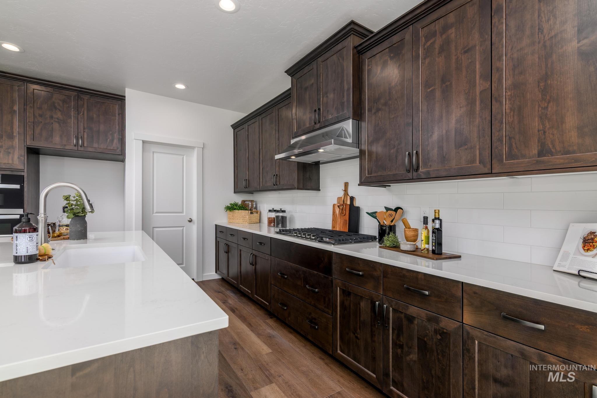 Kitchen featuring dark brown cabinetry, light stone counters, backsplash, dark wood-style floors, and under cabinet range hood