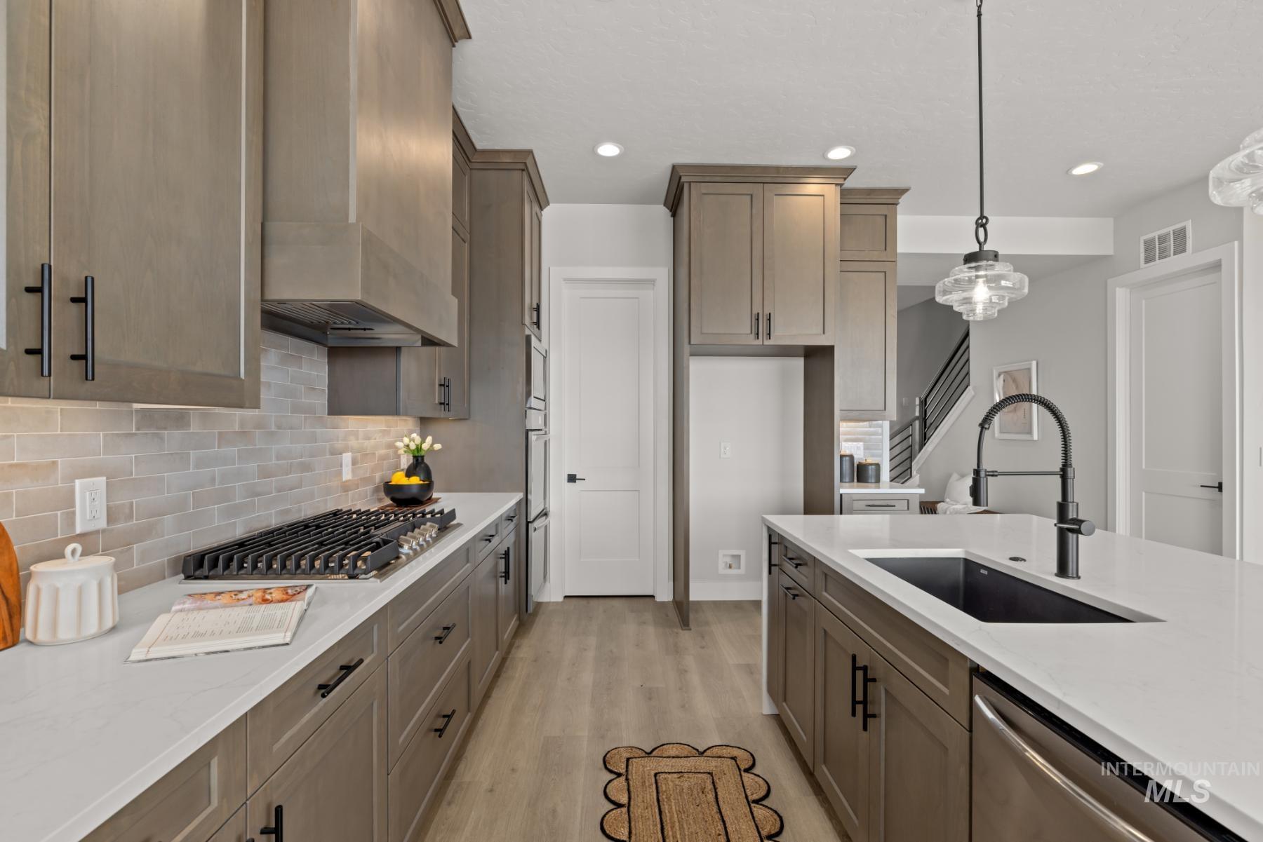 Kitchen with light stone counters, light wood-type flooring, hanging light fixtures, custom exhaust hood, and backsplash