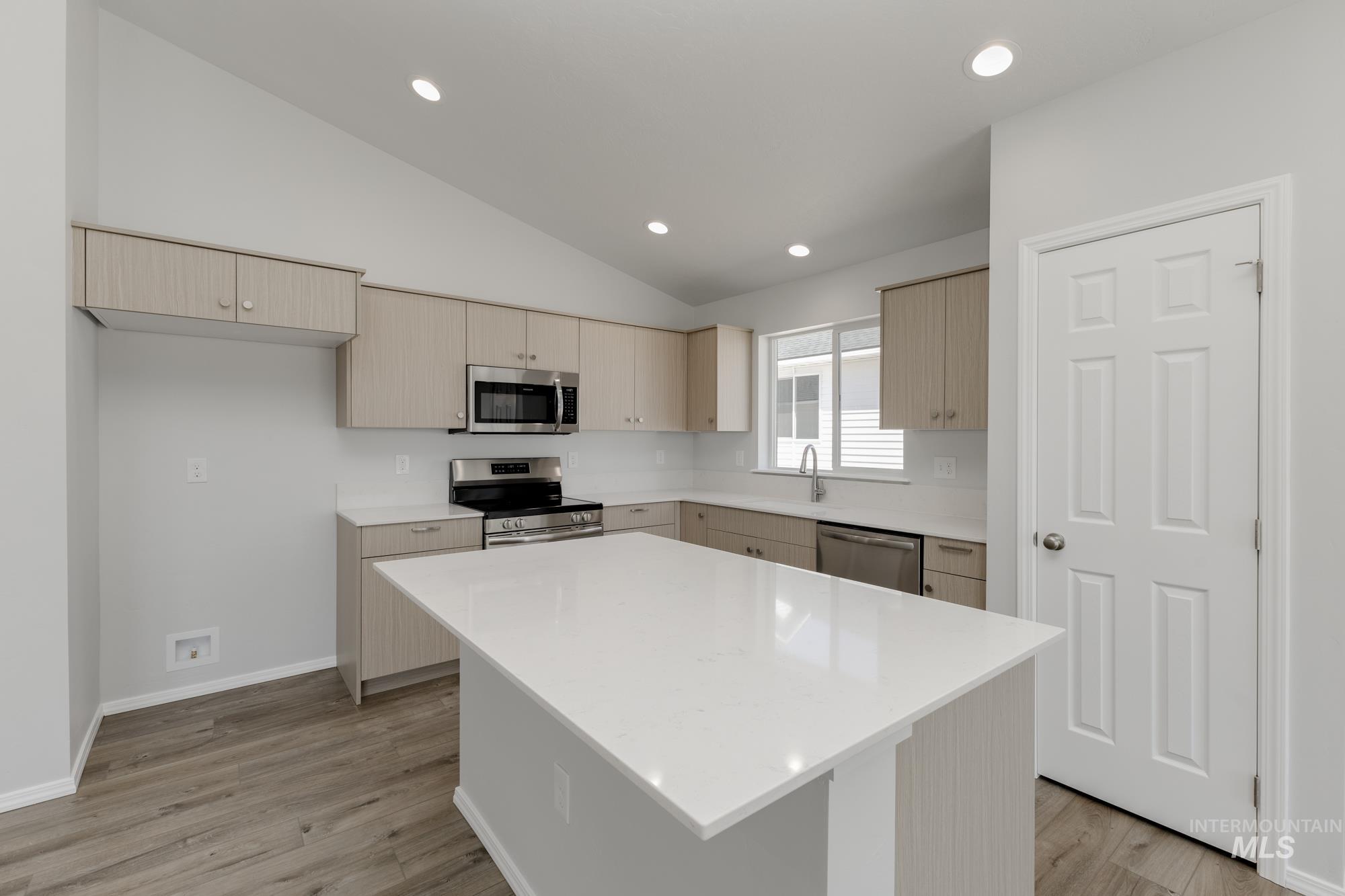 Kitchen featuring stainless steel appliances, a center island, light stone countertops, light brown cabinets, and vaulted ceiling