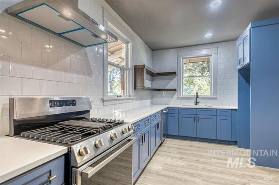 Kitchen featuring stainless steel appliances, range hood, decorative backsplash, blue cabinets, and light wood-style flooring
