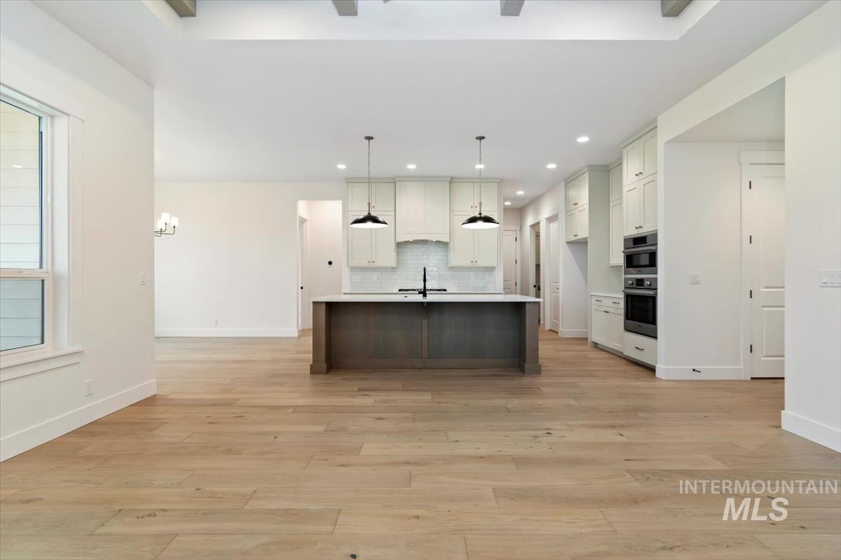 Kitchen featuring a kitchen island with sink, white cabinetry, decorative backsplash, hanging light fixtures, and premium range hood