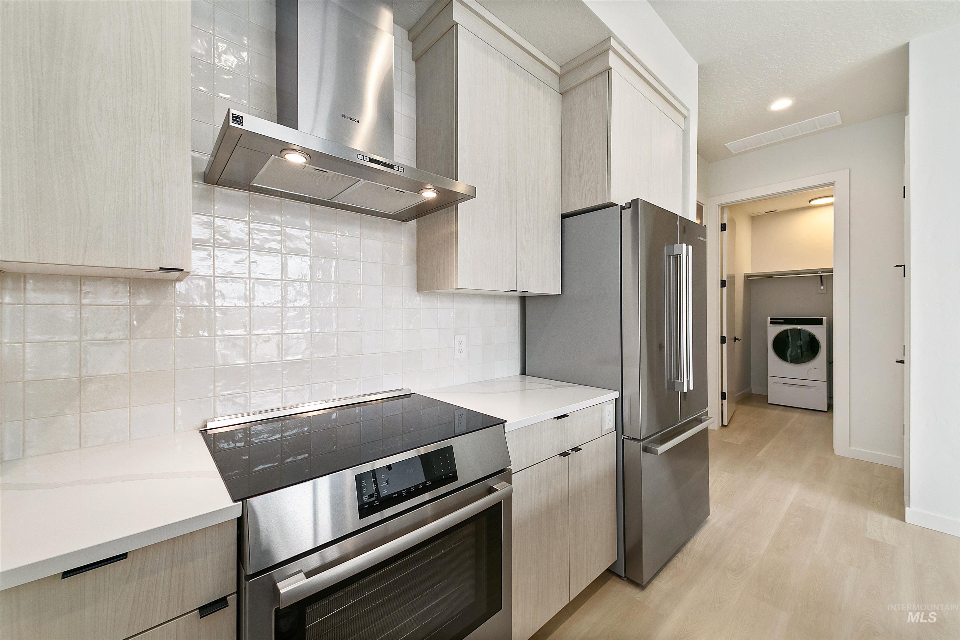 Kitchen featuring wall chimney exhaust hood, appliances with stainless steel finishes, washer / dryer, light wood-style flooring, and light brown cabinets