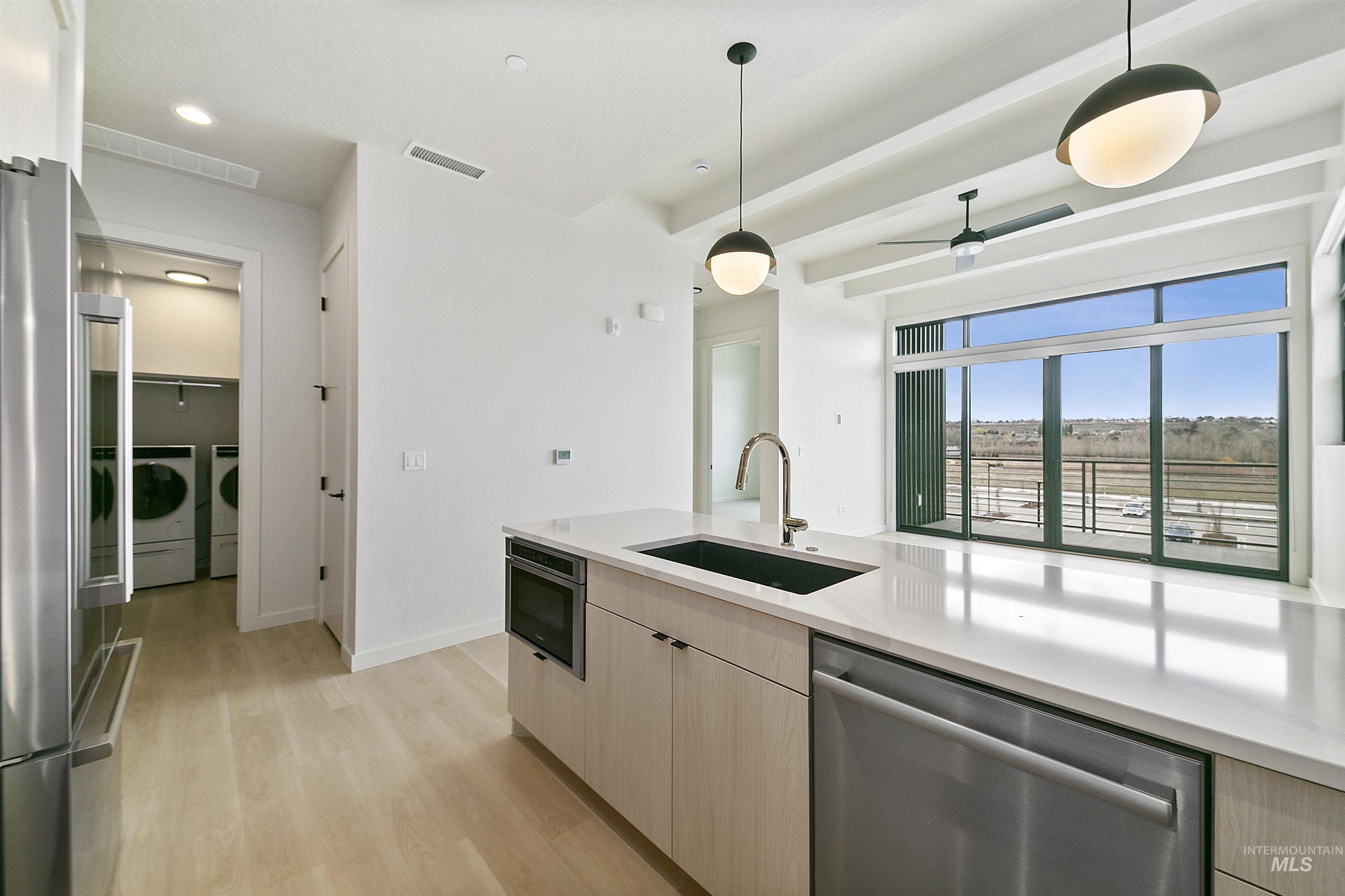 Kitchen with stainless steel appliances, light brown cabinetry, hanging light fixtures, light wood-style flooring, and beamed ceiling
