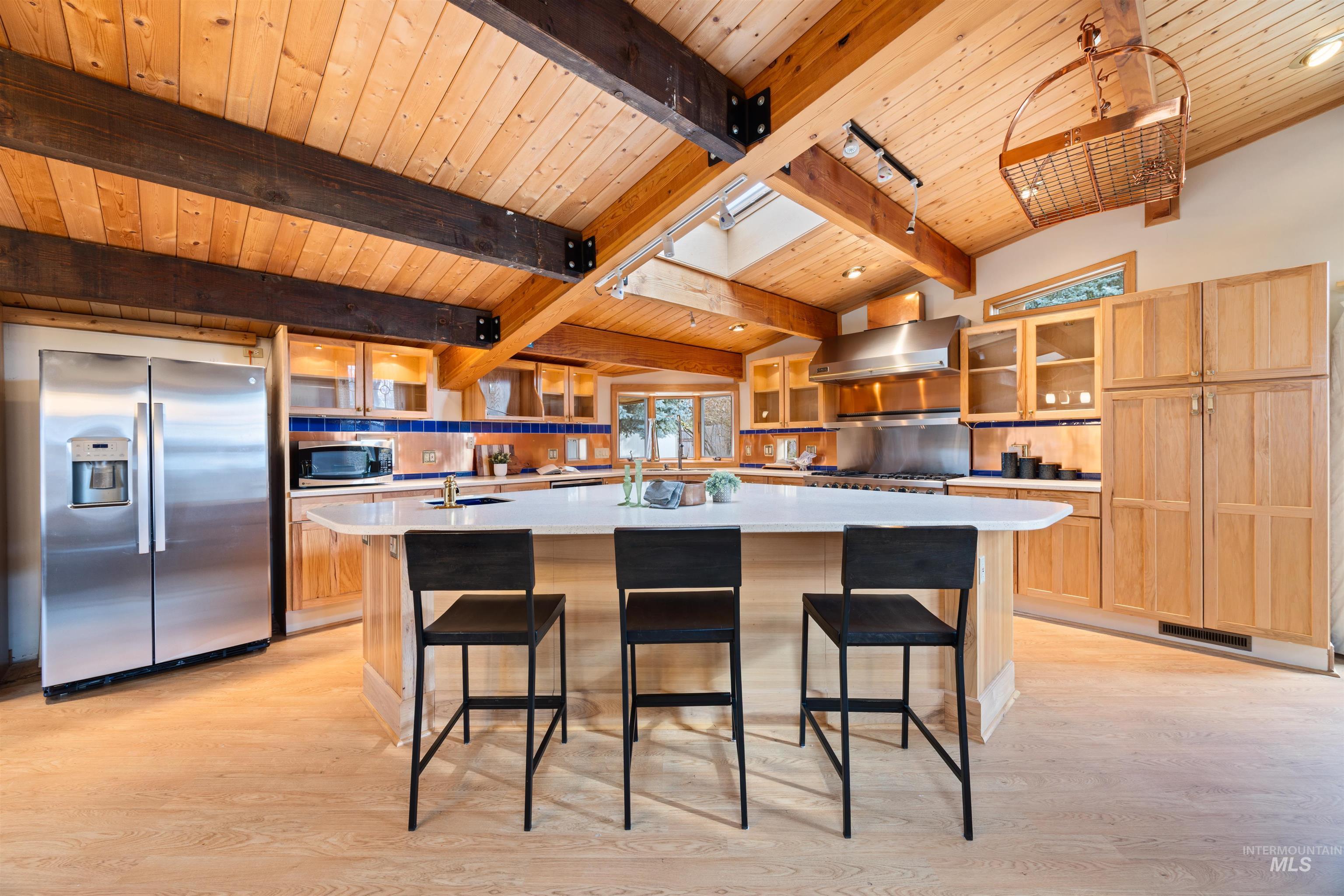 Kitchen featuring glass insert cabinets, a wooden ceiling with exposed beams, appliances with stainless steel finishes, a center island with sink, and wall chimney range hood