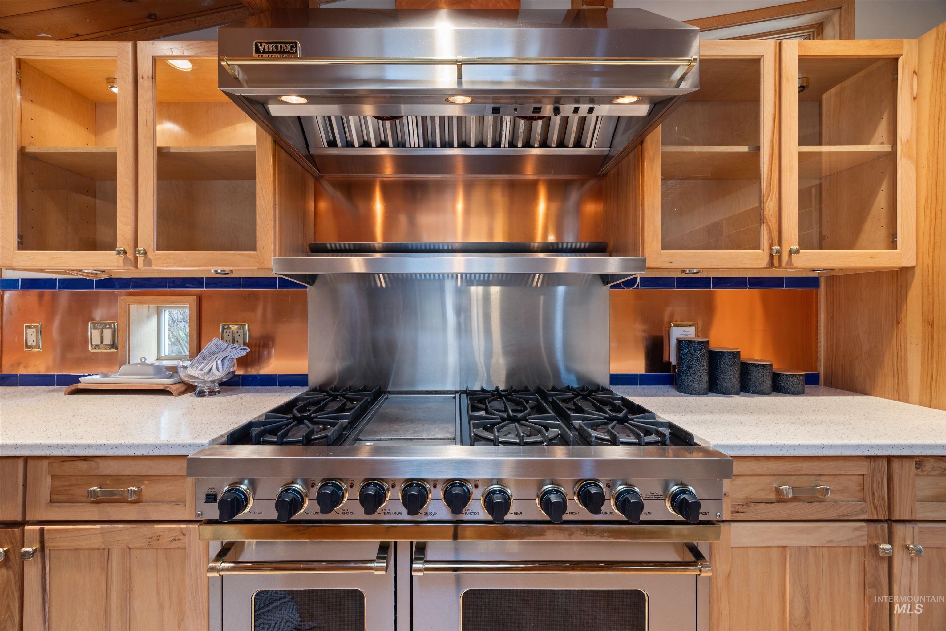 Kitchen featuring exhaust hood, glass insert cabinets, range with two ovens, backsplash, and light stone counters