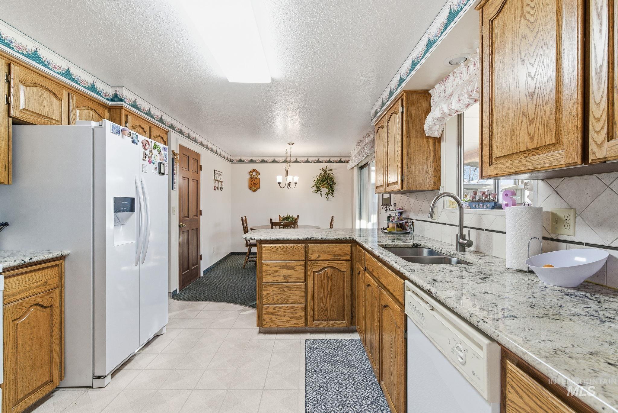 Kitchen with white appliances, a peninsula, brown cabinetry, light stone countertops, and hanging light fixtures
