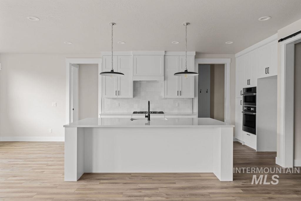 Kitchen featuring white cabinetry, pendant lighting, an island with sink, decorative backsplash, and light wood finished floors