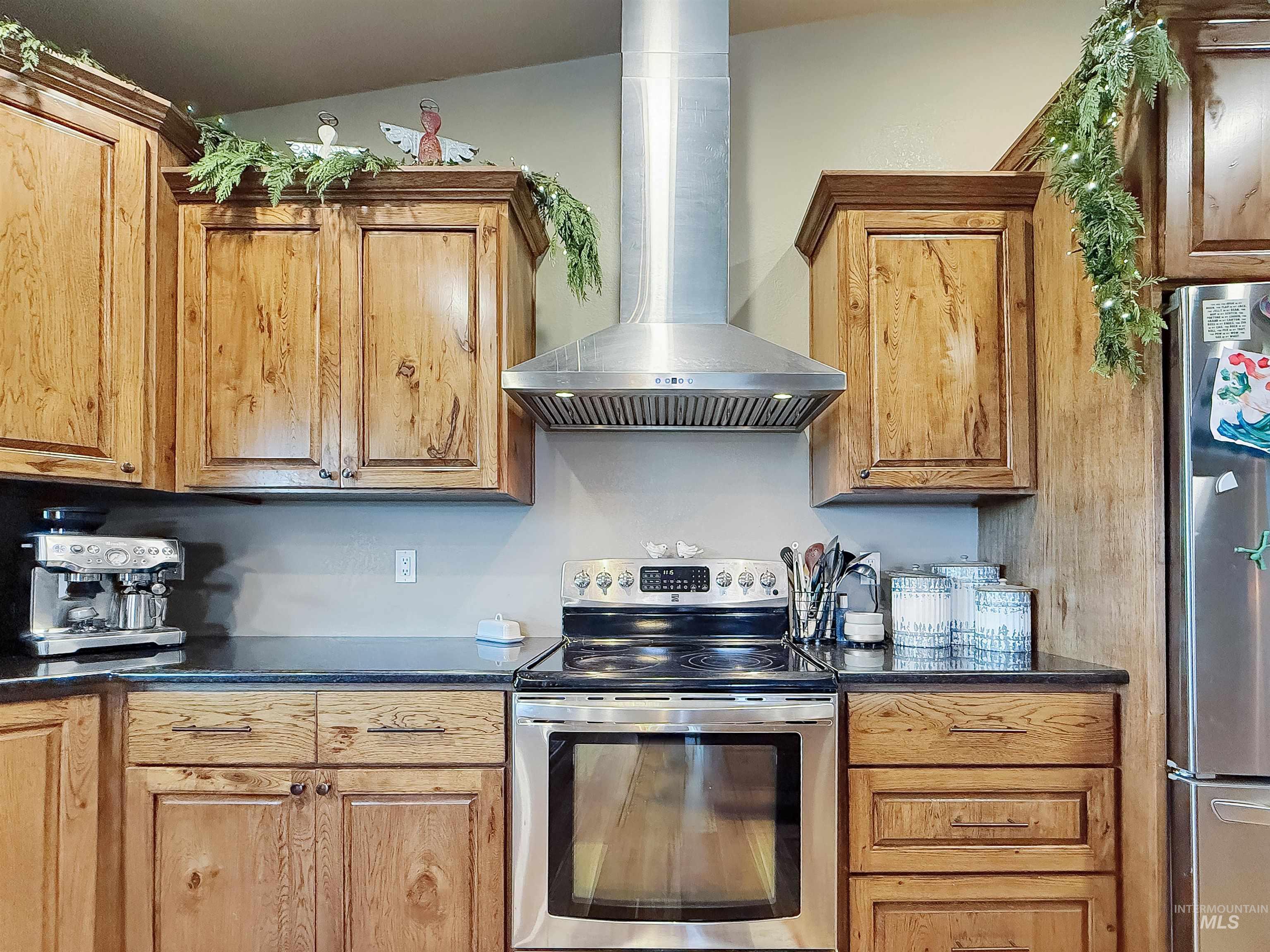 Kitchen with appliances with stainless steel finishes, wall chimney exhaust hood, dark stone countertops, and brown cabinets
